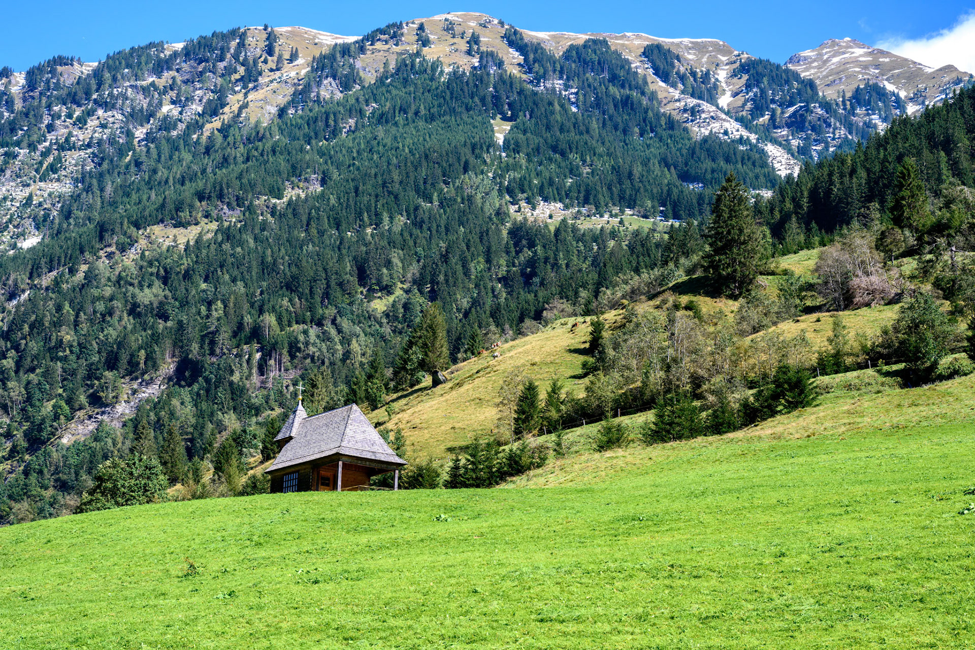 A wooden chapel stands on a hill on a green meadow in front of a mountain massif.