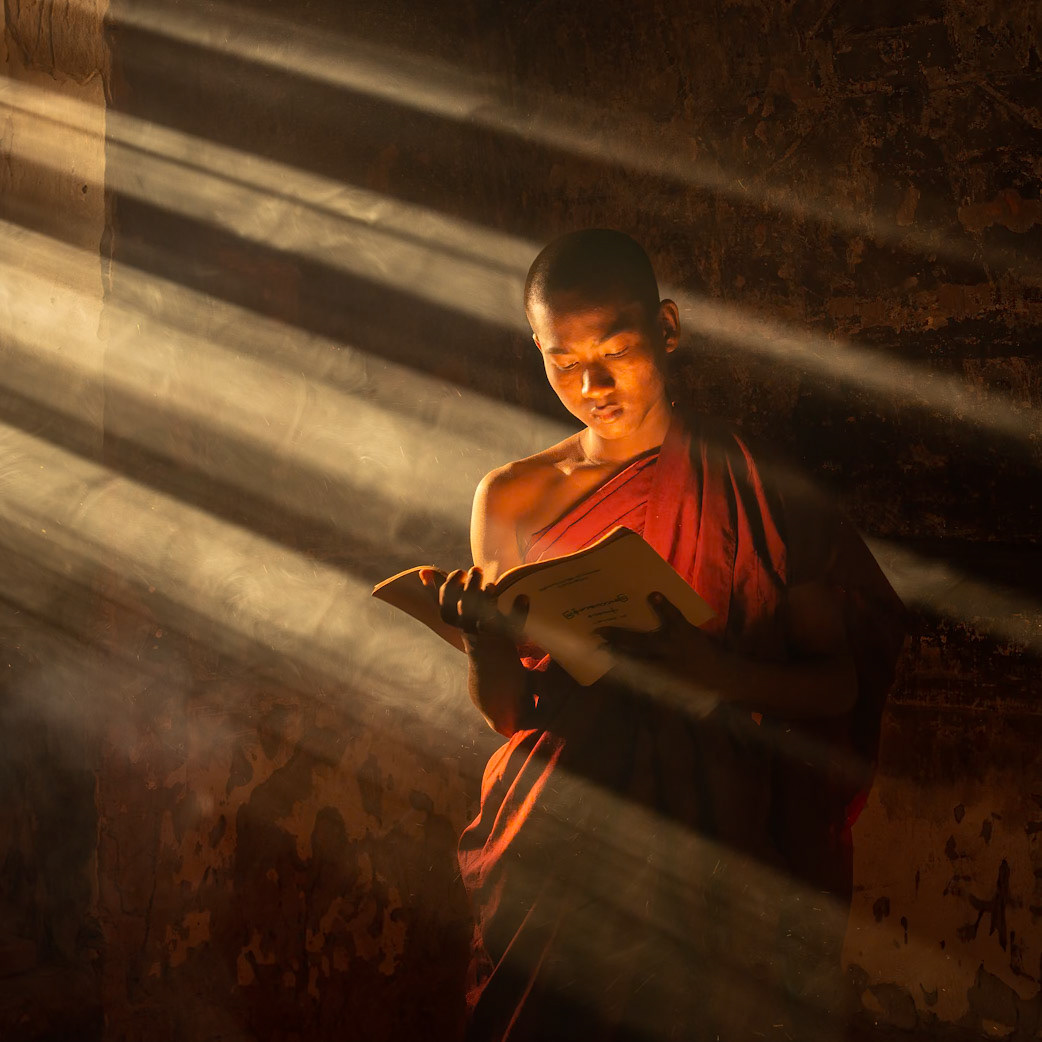 The streams of sunlight became rays of a higher kind as they lit their way through the smoky incence. The light lit the edge of the prayer book  while the reflecting light from the prayer book illuminated the young monks face.  It was as if the words of the prayer book illumniated the young monks face. .