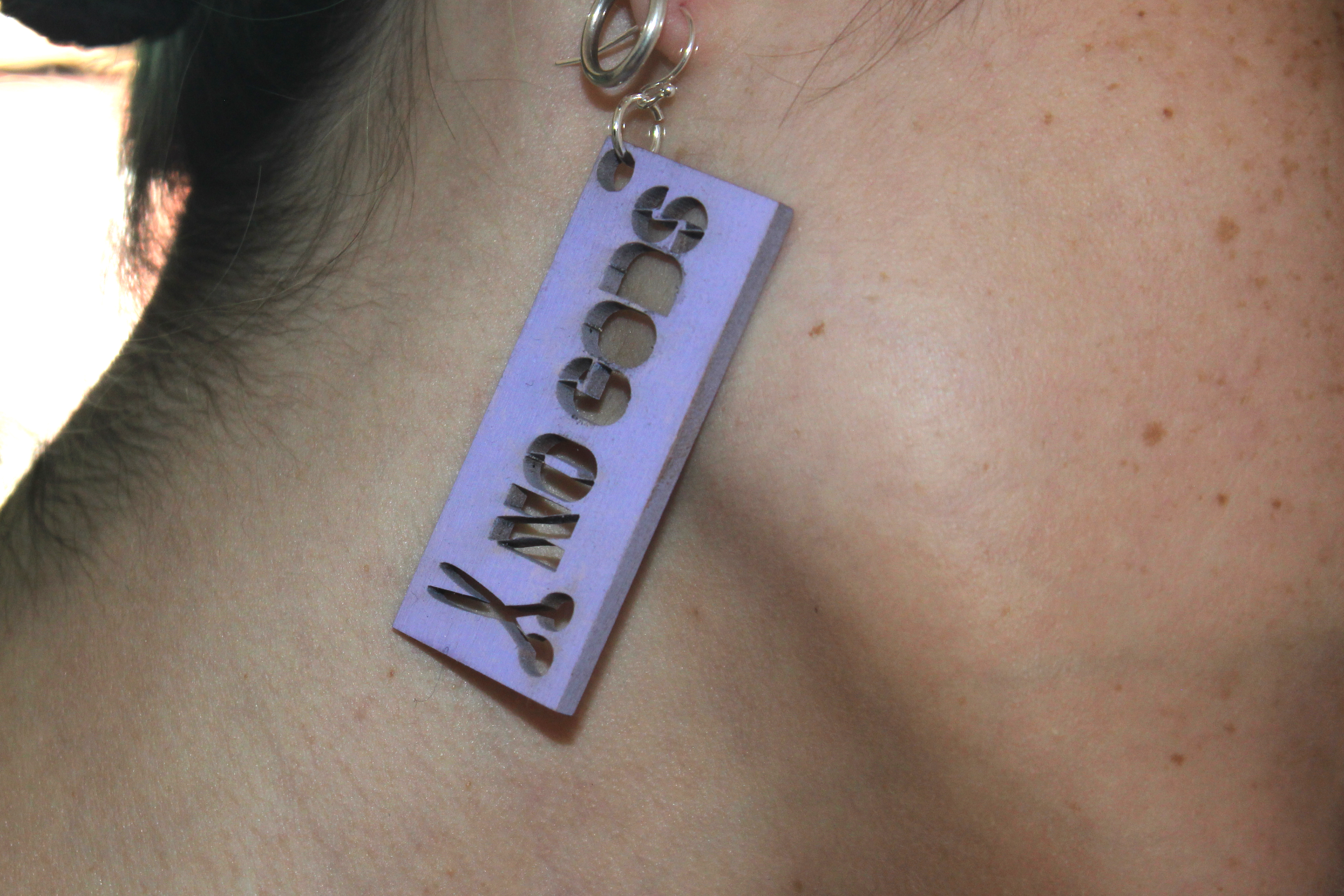 a wood stencil earring hanging from an ear. it says "no gods" and has a small pair of scissors on it.