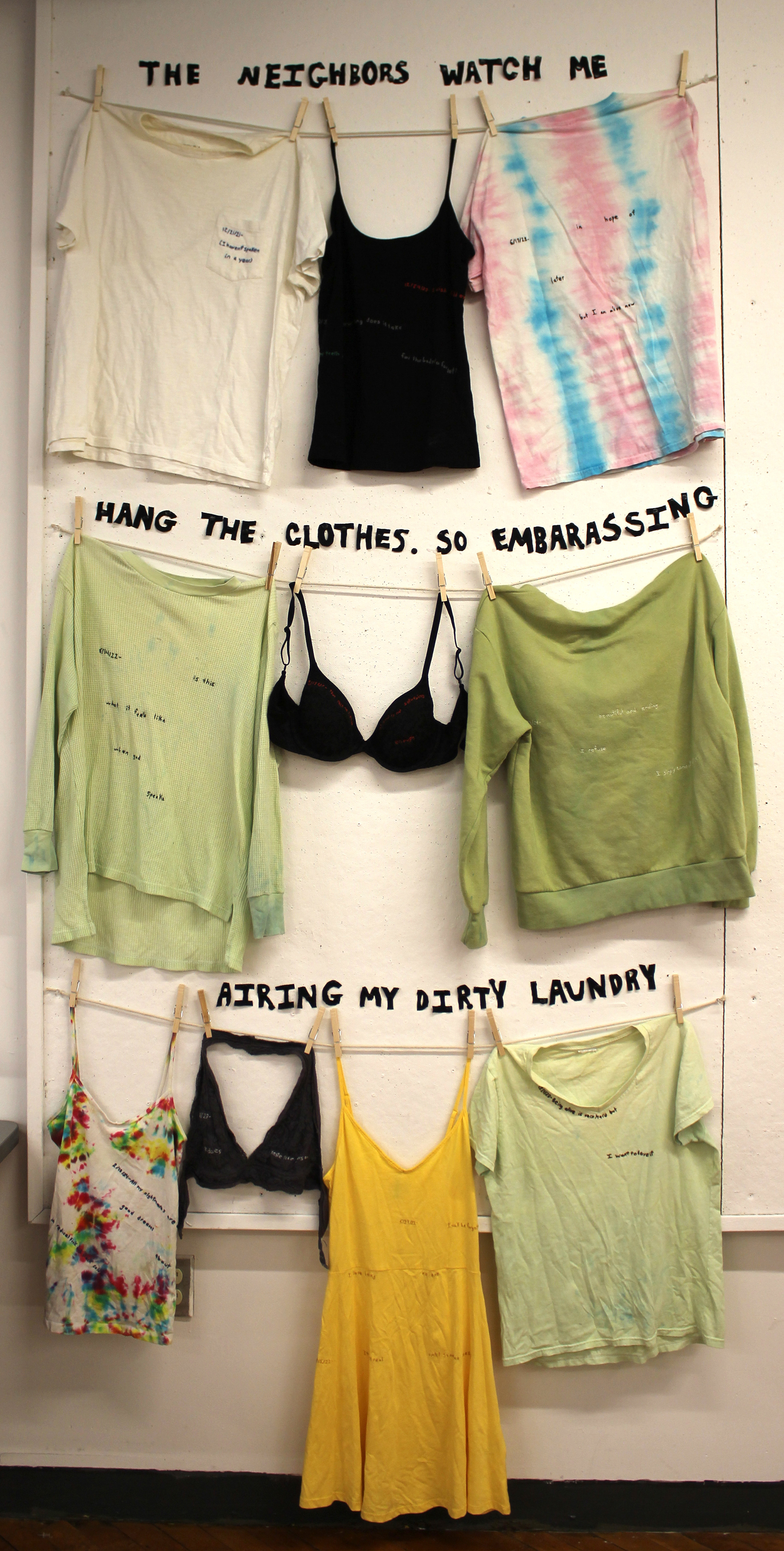 instillation featuring three tiers of clothes hanging on a clothesline. Black text made of cut up pieces of clothes is pinned to the wall reading "the neighbors watch me hang the clothes. So embarrassing airing my dirty laundry"