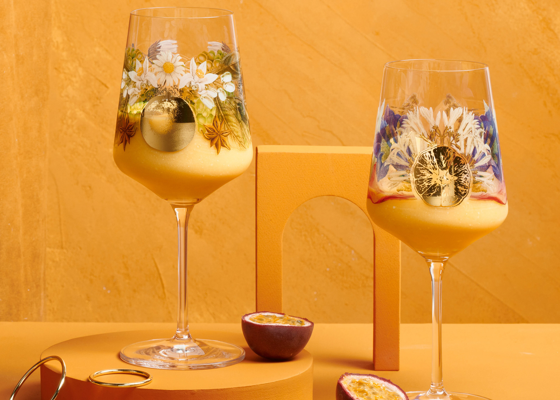 A photo of 2 long stem aperitif glasses with floral imagery on the glass against an orange background.