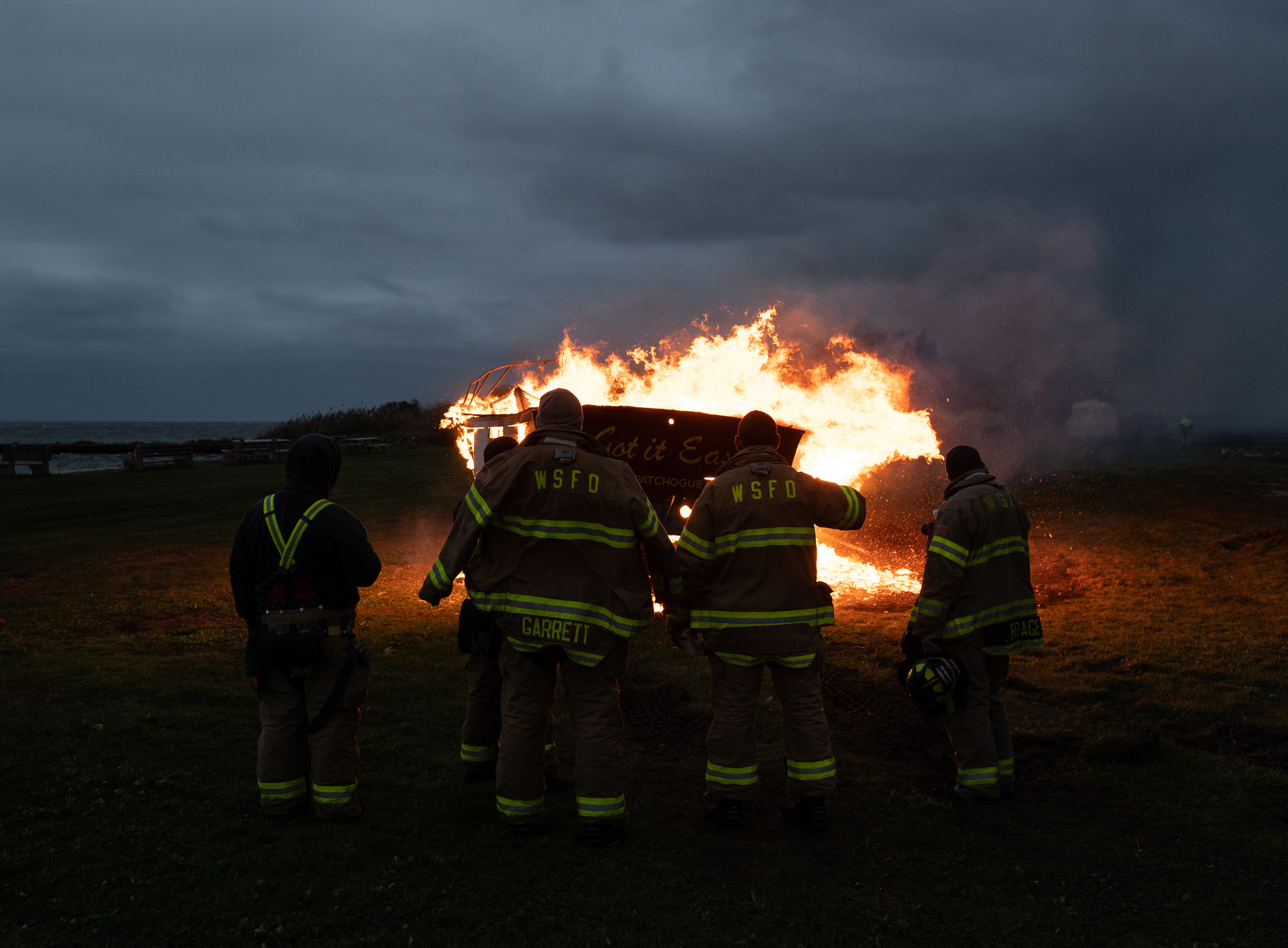Several firefighters representing the West Sayville Fire Department look on in awe at a burning boat during the Long Island Maritime Museum’s 2021 Halloween Boat Burning event.