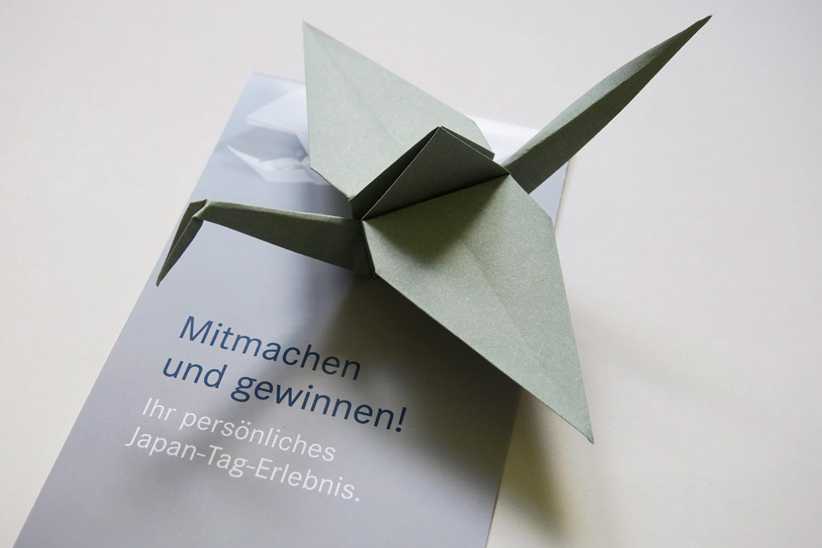 Folded origami crane and flyer with folding instructions for a customer to distribute at a trade fair stand.