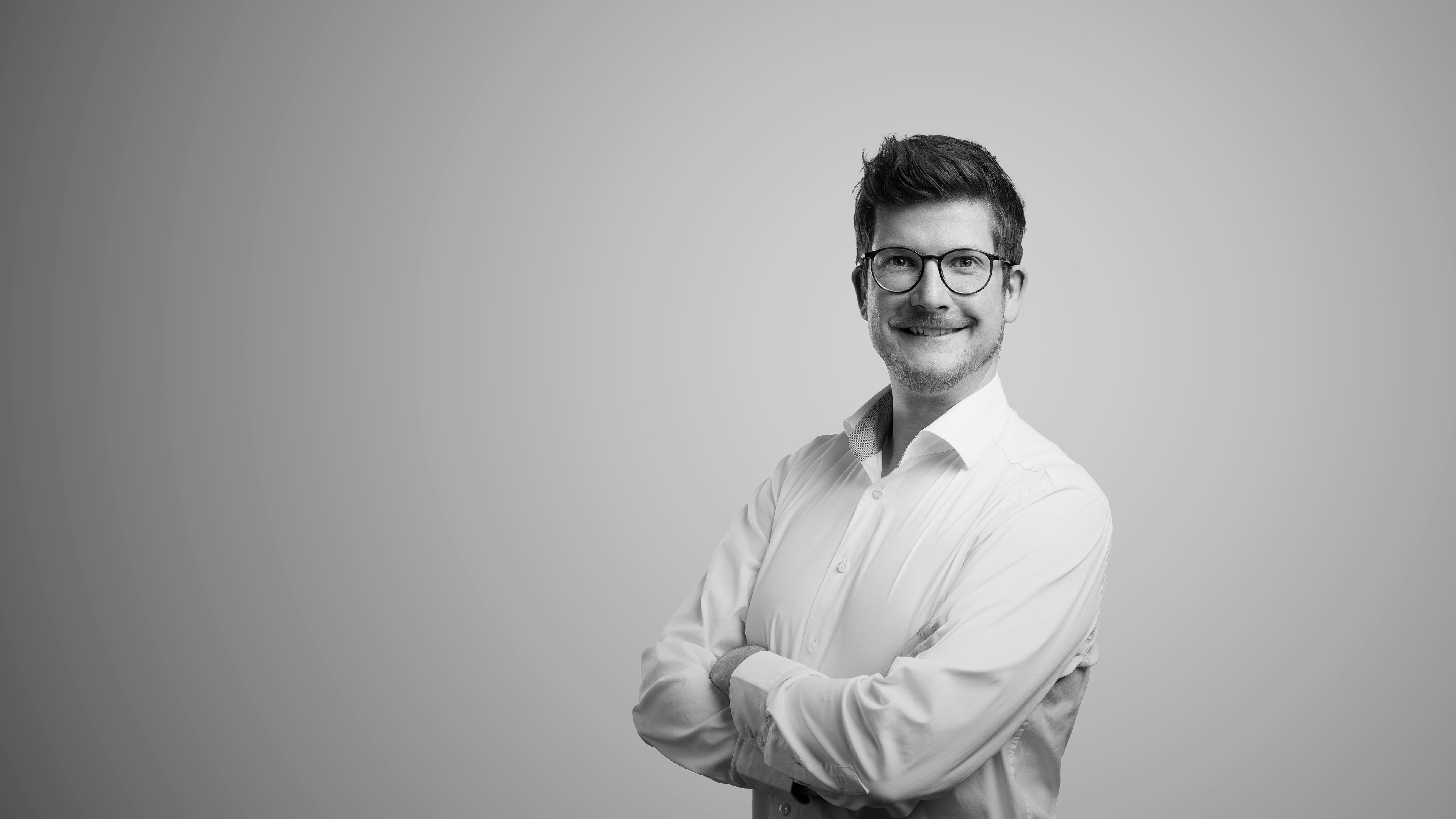 Schwarz-weiß-Porträt von Marcel Cornelius. Er trägt eine Brille und ein weißes Hemd, steht mit verschränkten Armen vor einem neutralen, grauen Hintergrund und lächelt selbstbewusst in die Kamera.