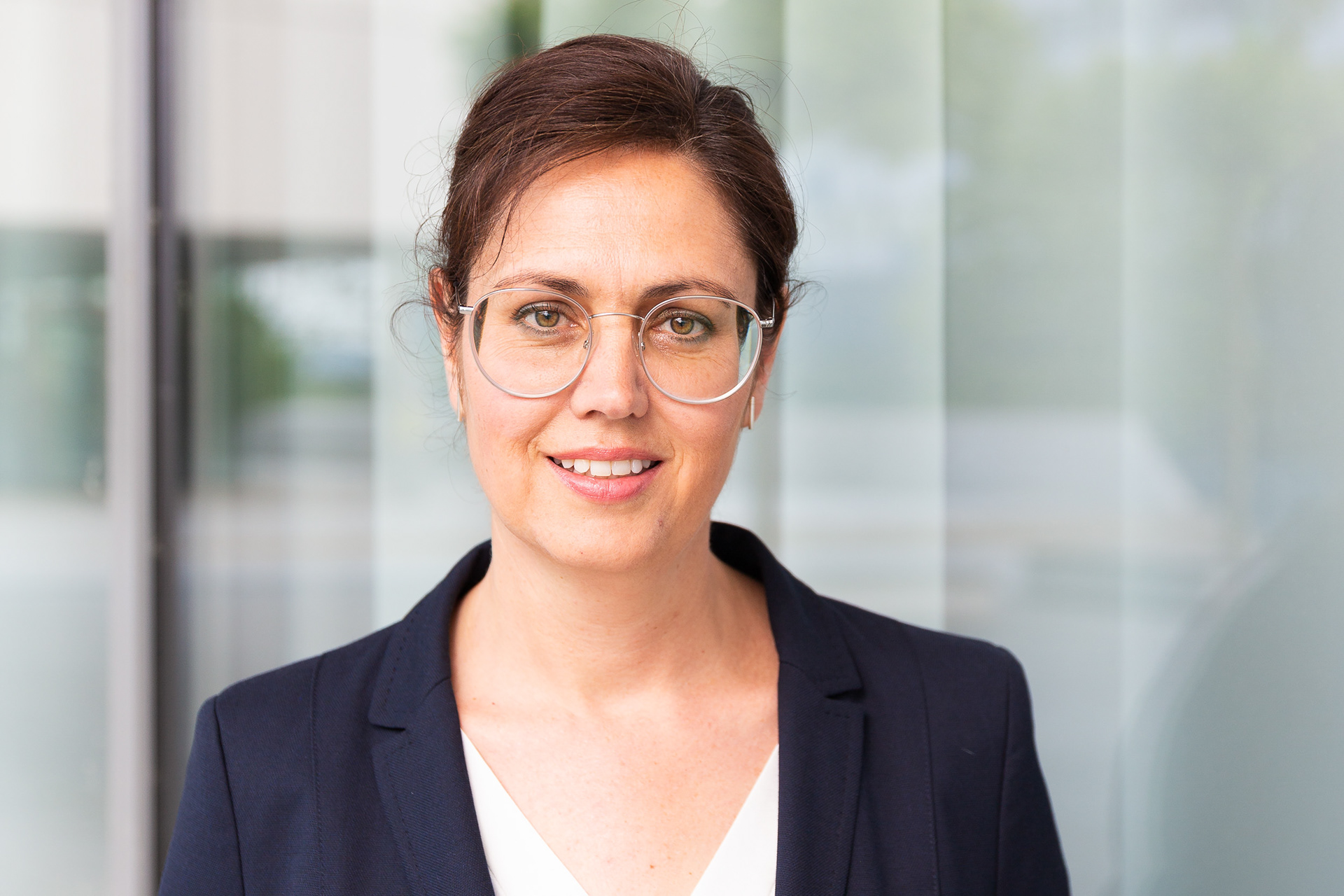 Porträt von Ruth Meyhöfer, Geschäftsführerin der Perspect GmbH. Sie trägt eine runde Brille und einen dunkelblauen Blazer über einer weißen Bluse. Der Hintergrund zeigt eine moderne Glasfassade mit sanften Spiegelungen.