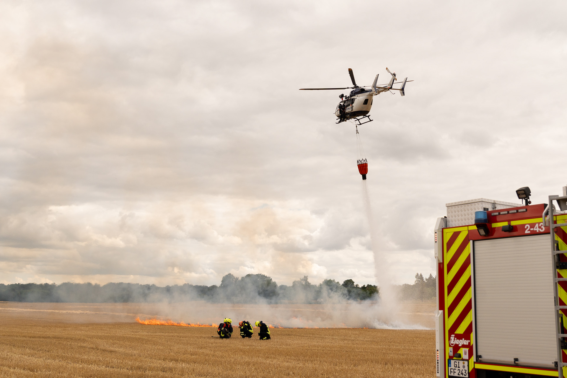 Polizeihubschrauber beim Löschwasserabwurf über einem brennenden Feld während der Vegetationsbrandübung ‚Inferno Experts – Annerod 2025‘, im Vordergrund Einsatzkräfte der Feuerwehr Fernwald und ein Löschfahrzeug.
