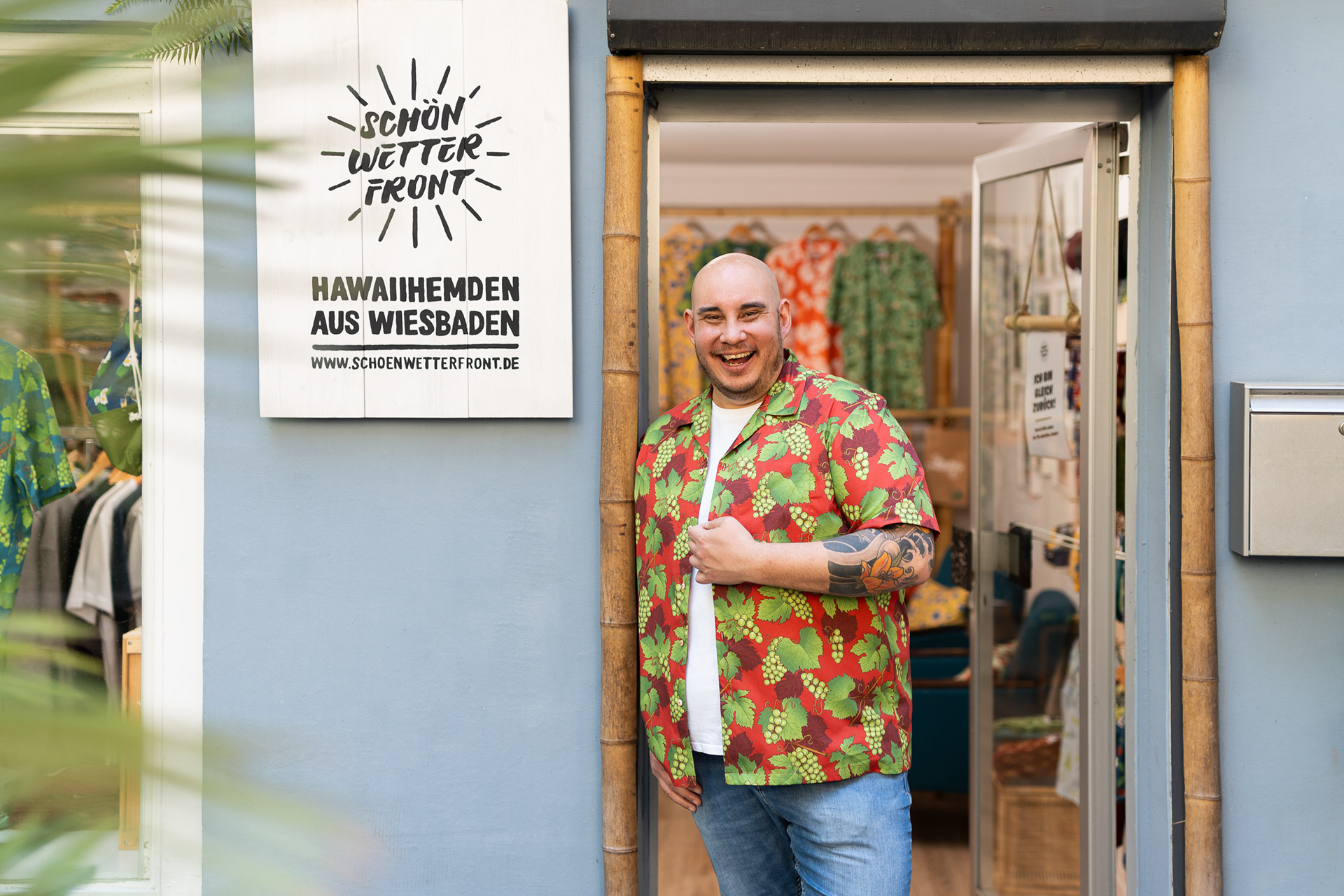 Christian von Schönwetterfront steht lächelnd in der Tür seines Ladens in Wiesbaden. Er trägt ein auffälliges rotes Hawaiihemd mit grünem Traubenmuster und eine Jeans. Sein rechter Arm ist tätowiert. Links neben ihm hängt ein weißes Holzschild mit dem Schriftzug ‚Schönwetterfront – Hawaiihemden aus Wiesbaden‘ und der Website-Adresse. Im Hintergrund sind farbenfrohe Hawaiihemden auf Kleiderbügeln zu sehen.