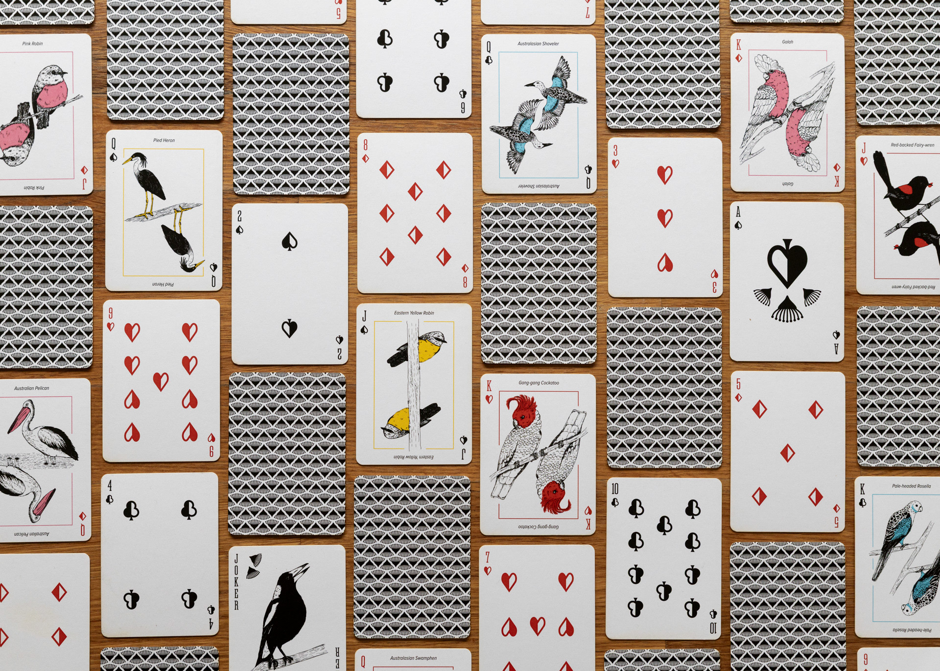 A photo of the the playing cards laid out on a wooden table, taken from directly overhead. The cards are arranged in a vertical brick pattern. SOme cards are face up, and others are face down, showing the repeating gum blossom pattern on the reverse.