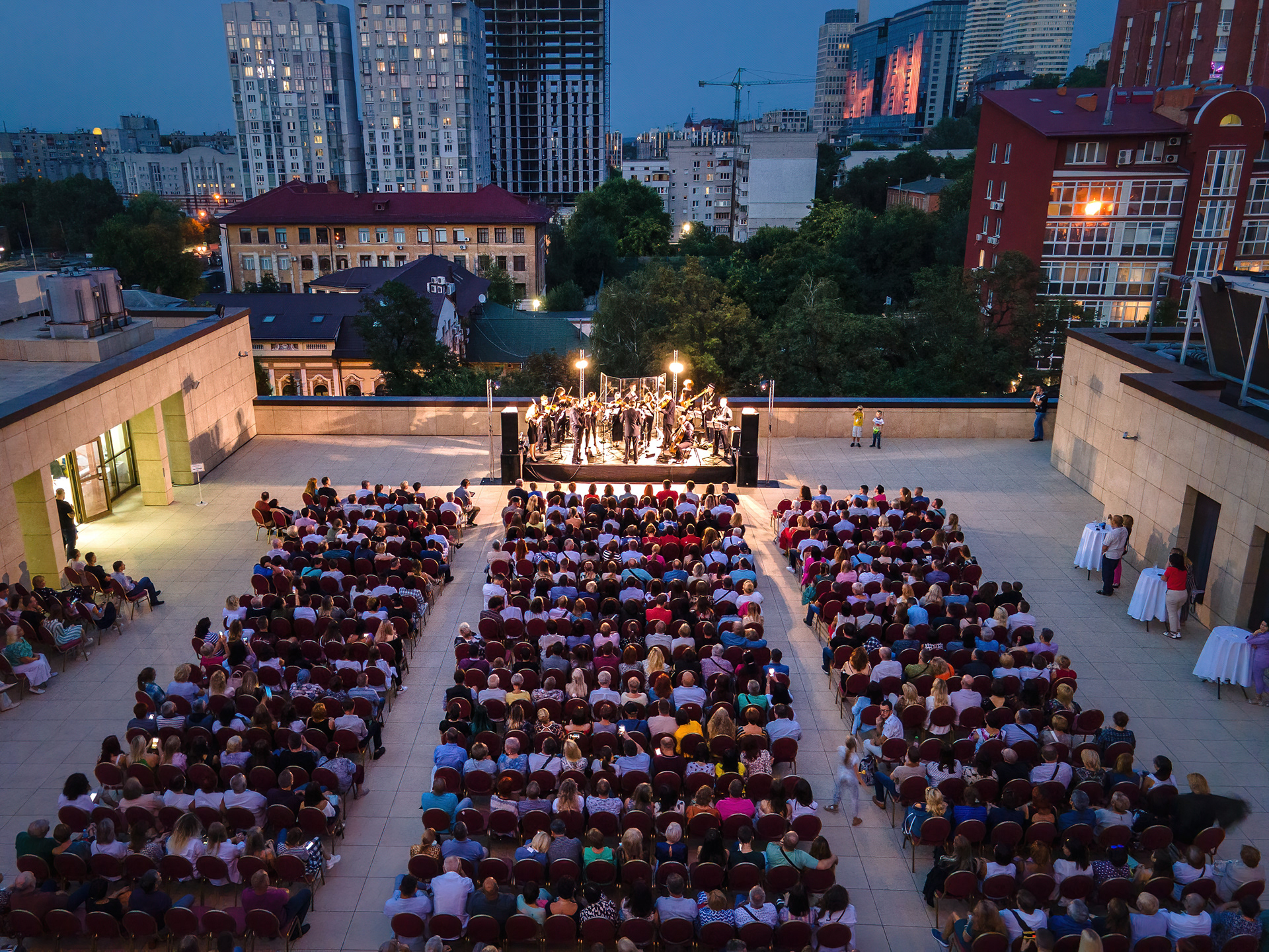 Evening rooftop concert from above, a full audience facing the lit stage