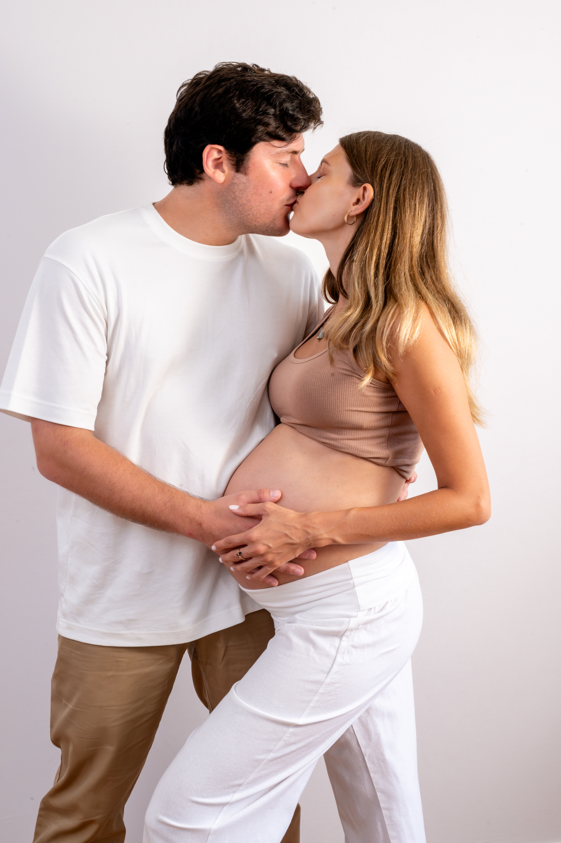 Pregnant couple kissing against a white wall