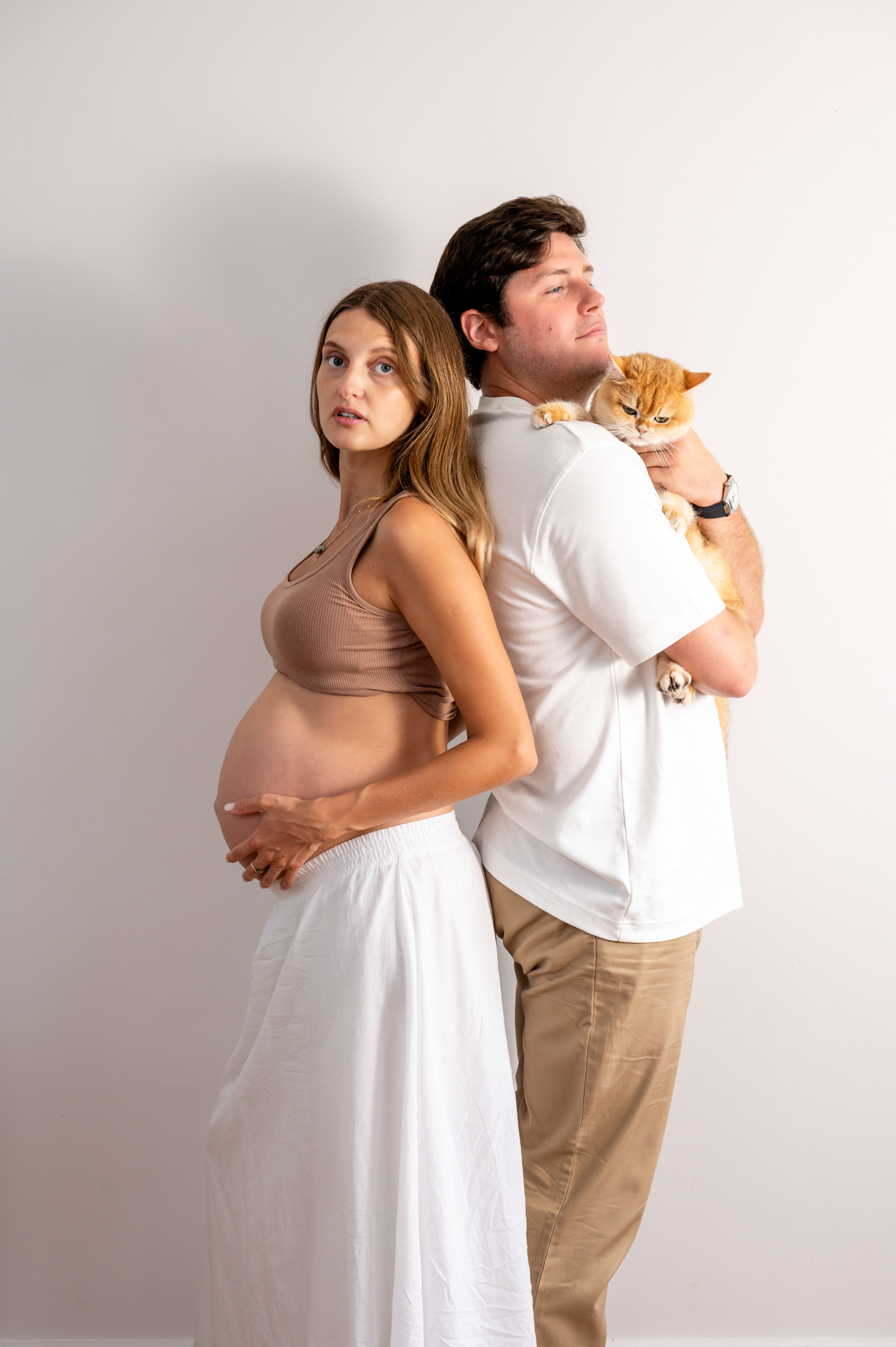 Expecting couple posing back to back with their ginger cat