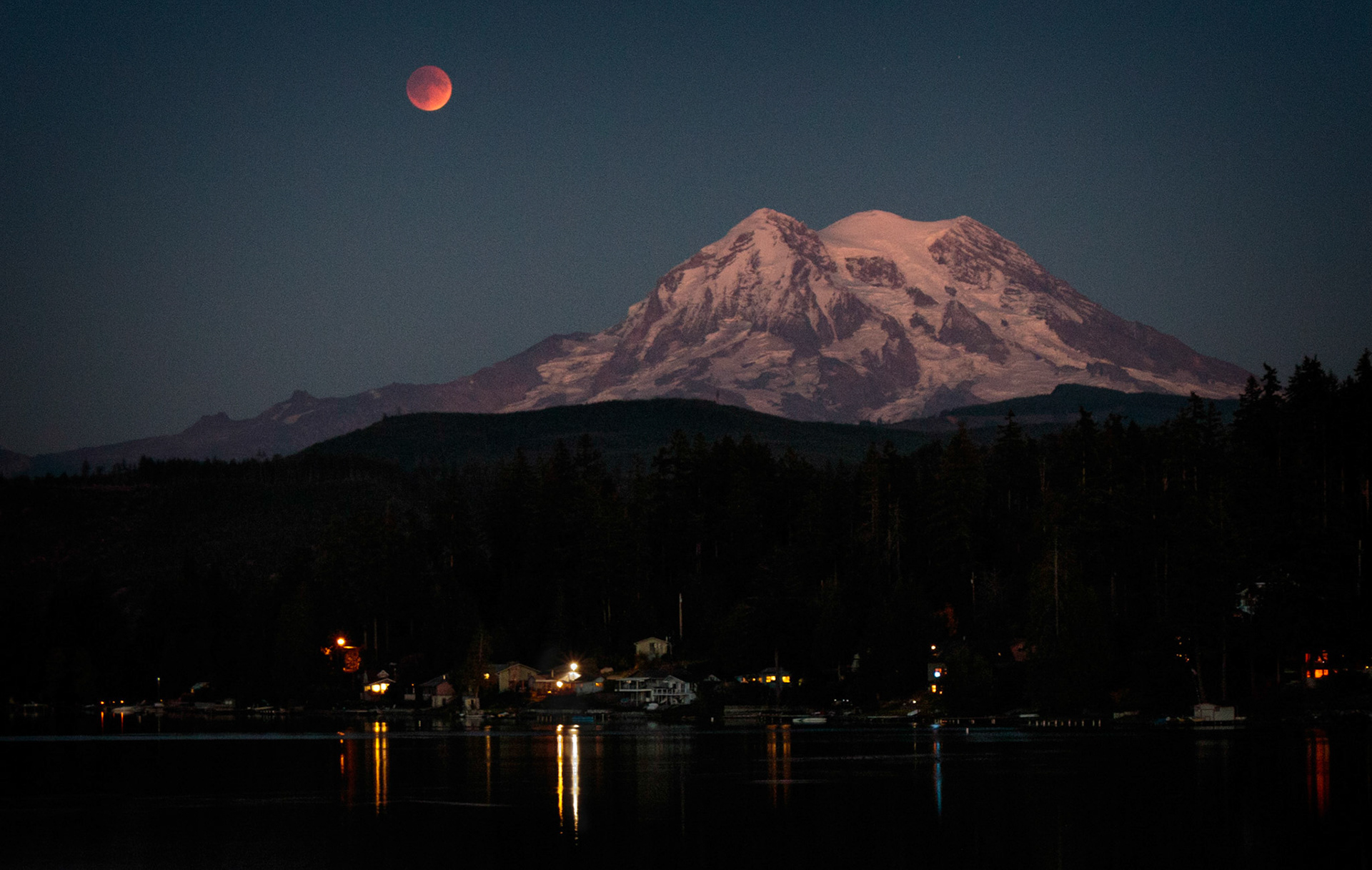The moon in total eclipse hangs blood red above Mt Rainier. The mountain and the moon are reflected in the waters of Clear Lake near Eatonville, WA.
