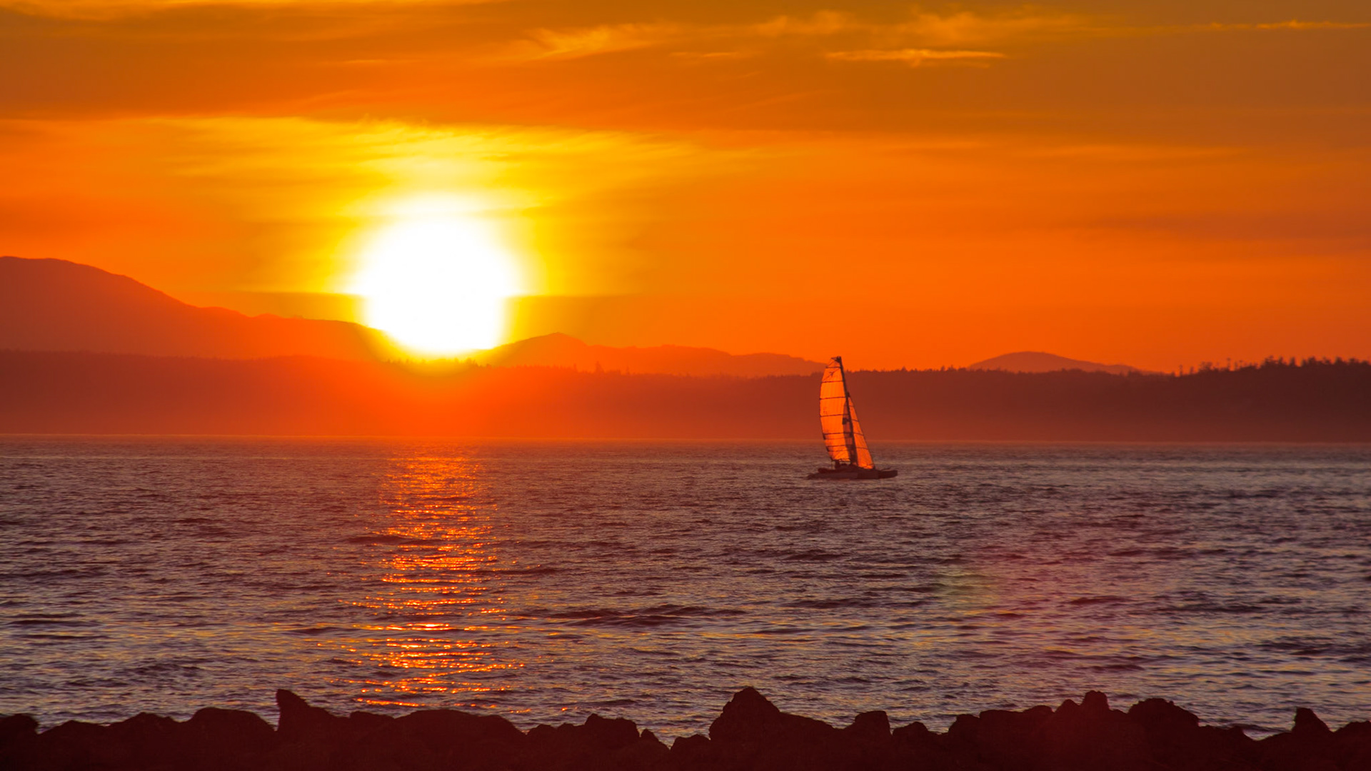 A sailboat reaches across Puget Sound as the sun is about to set behind the Olympic Mountains