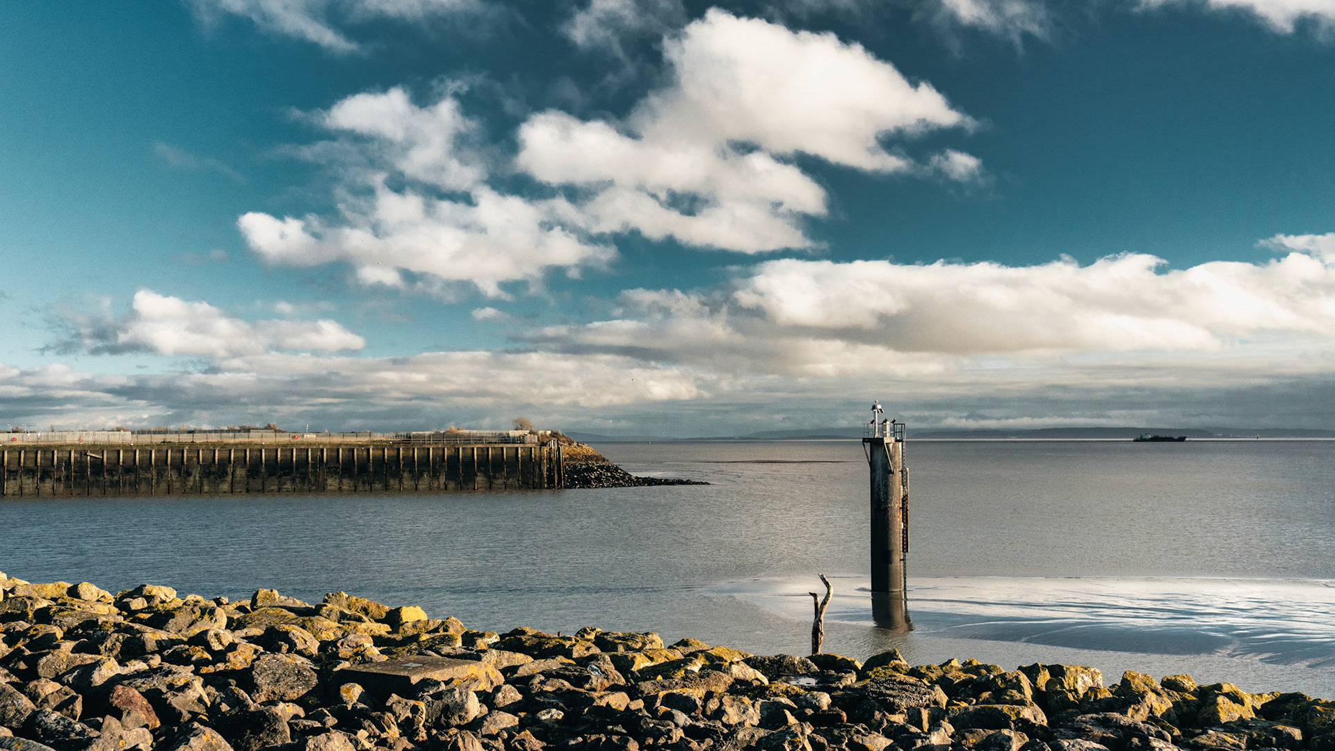 Waves whisper to the rocks as a lone beacon stands watch — a quiet sentinel beneath a sky painted with drifting clouds.