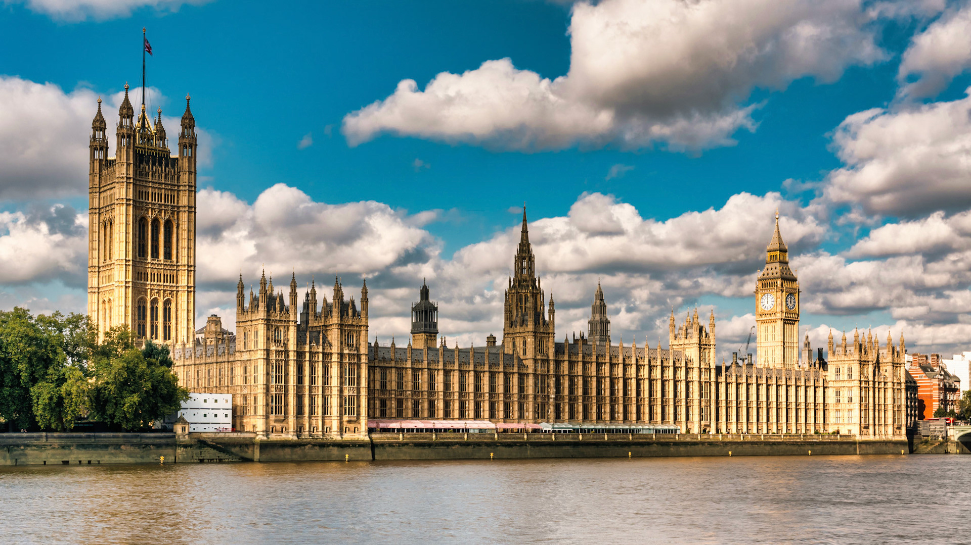 Bathed in golden sunlight, the iconic Palace of Westminster and Big Ben stand proudly against a canvas of clouds and blue skies along the River Thames.