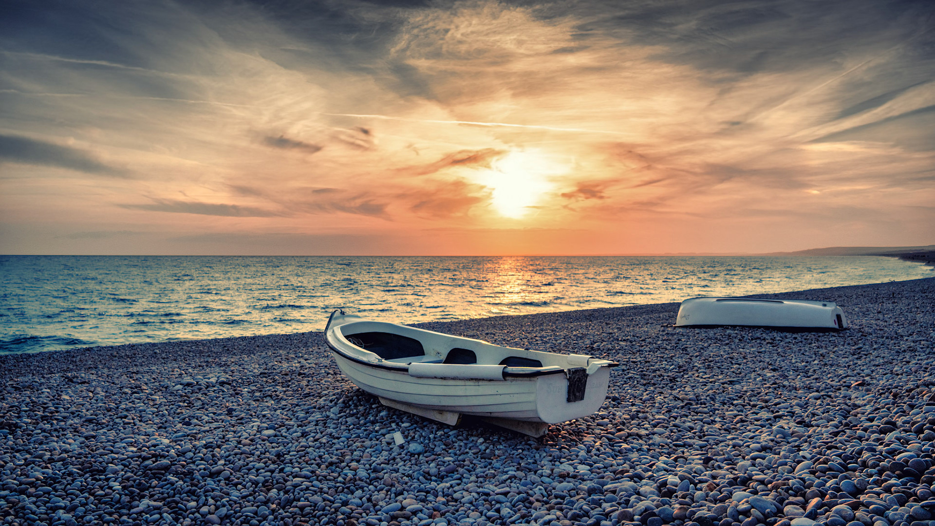 A quiet boat waits on the pebbled shores of Chesil Beach as the sun dips into the horizon, painting the sky with warmth.