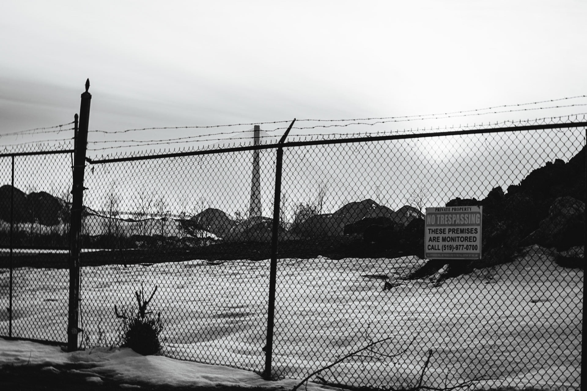 Monolithic Noir study of the Gordie Howe International Bridge pylon, rendered with maximum depth of field to include foreground industrial grit and fencing. A.M. Stone | Archival Stone Photography
