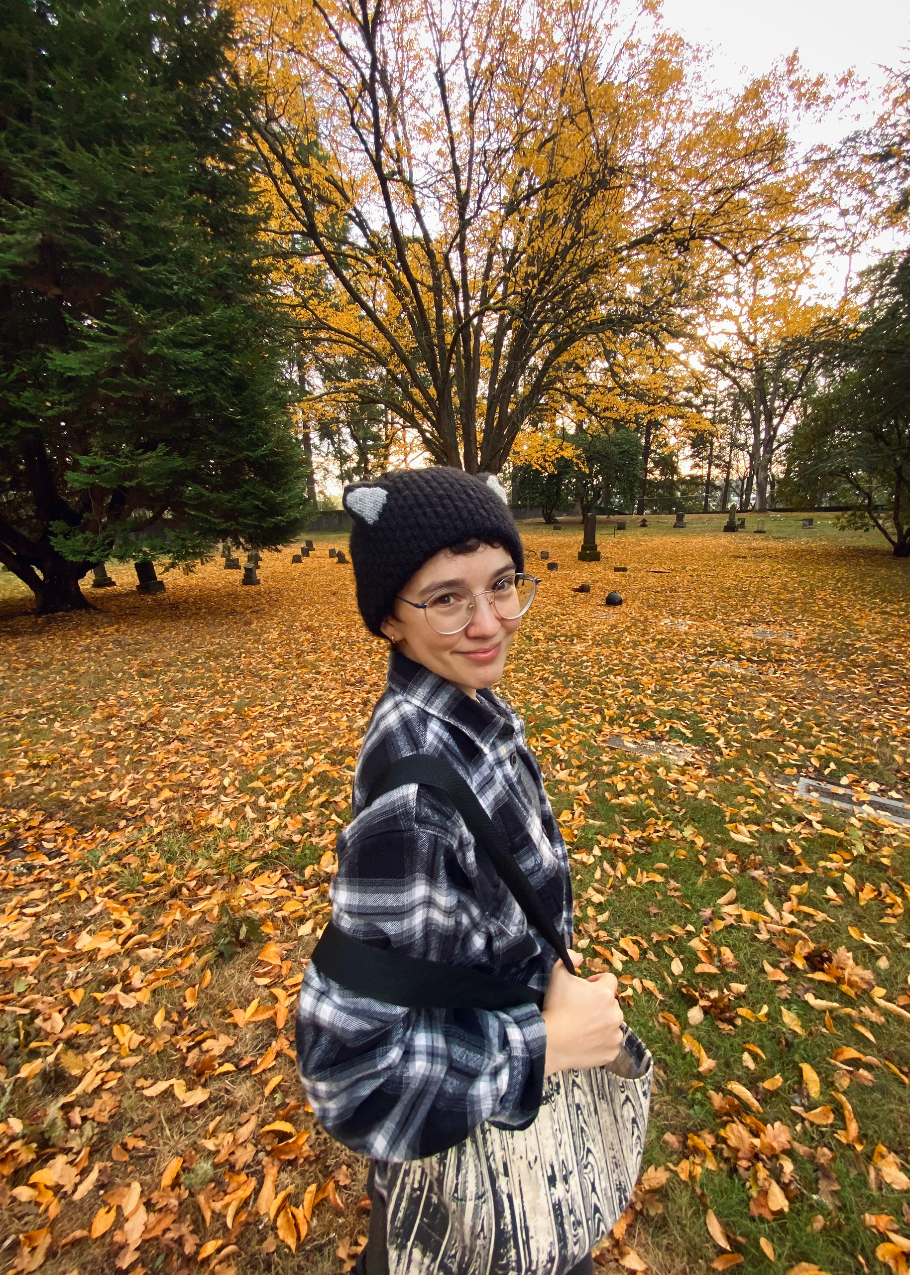 Alt Text: Sam, a white person in a black knit cat hat and black and white flannel, stands in front of a golden fall tree.