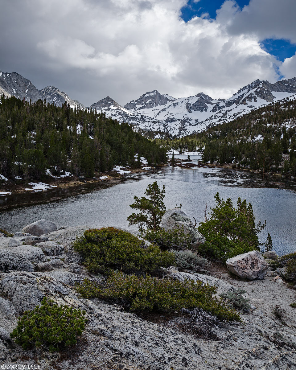 Shrubs grow on a rocky mound above Marsh Lake with the Sierra Nevada Mountains beyond in the John Muir Wilderness, California