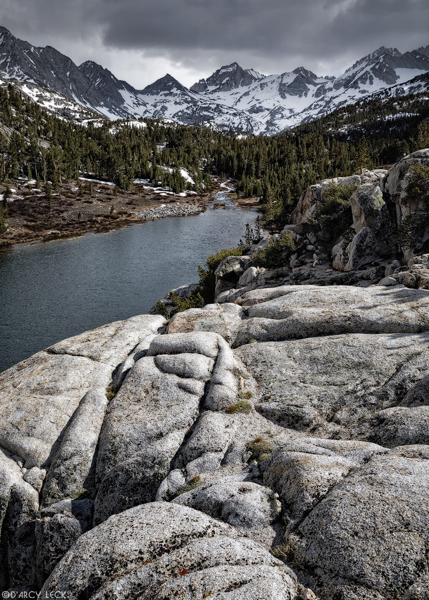 A rocky precipice overlooks Mack Lake with the Sierra Nevada Mountains beyond under late Spring snow showers in Little Lakes Valley, California