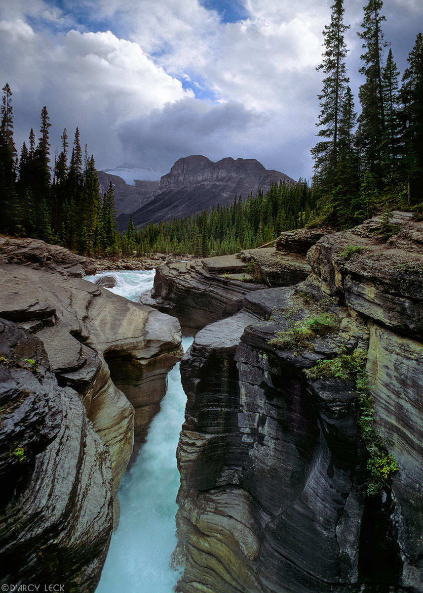 Mistaya River flows through Mistaya Canyon as late afternoon sunlight illuminates storm clouds over Kaufman Peaks, Alberta Canada