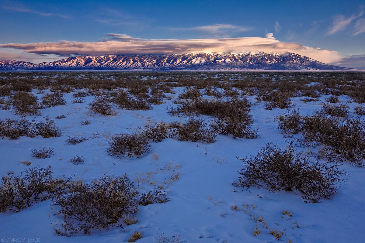 Lenticular clouds cap the snowy Blanca Peak and the Sangre de Cristo Mountains at sunset beyond the winter rangeland in the San Luis Valley, Colorado