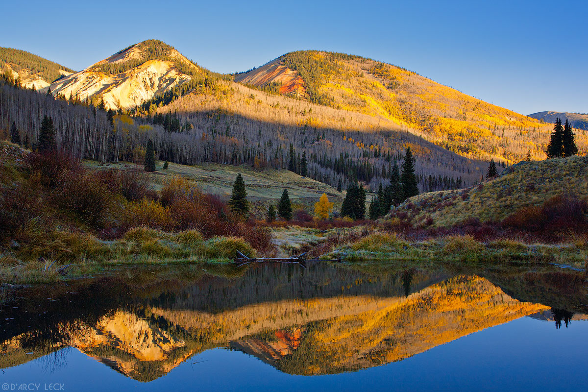 Mineral streaked Little Red Mountain catches the sun's rays at sunrise in autumn, and is reflected in a Beaver pond, Rio Grande National Forest, Colorado