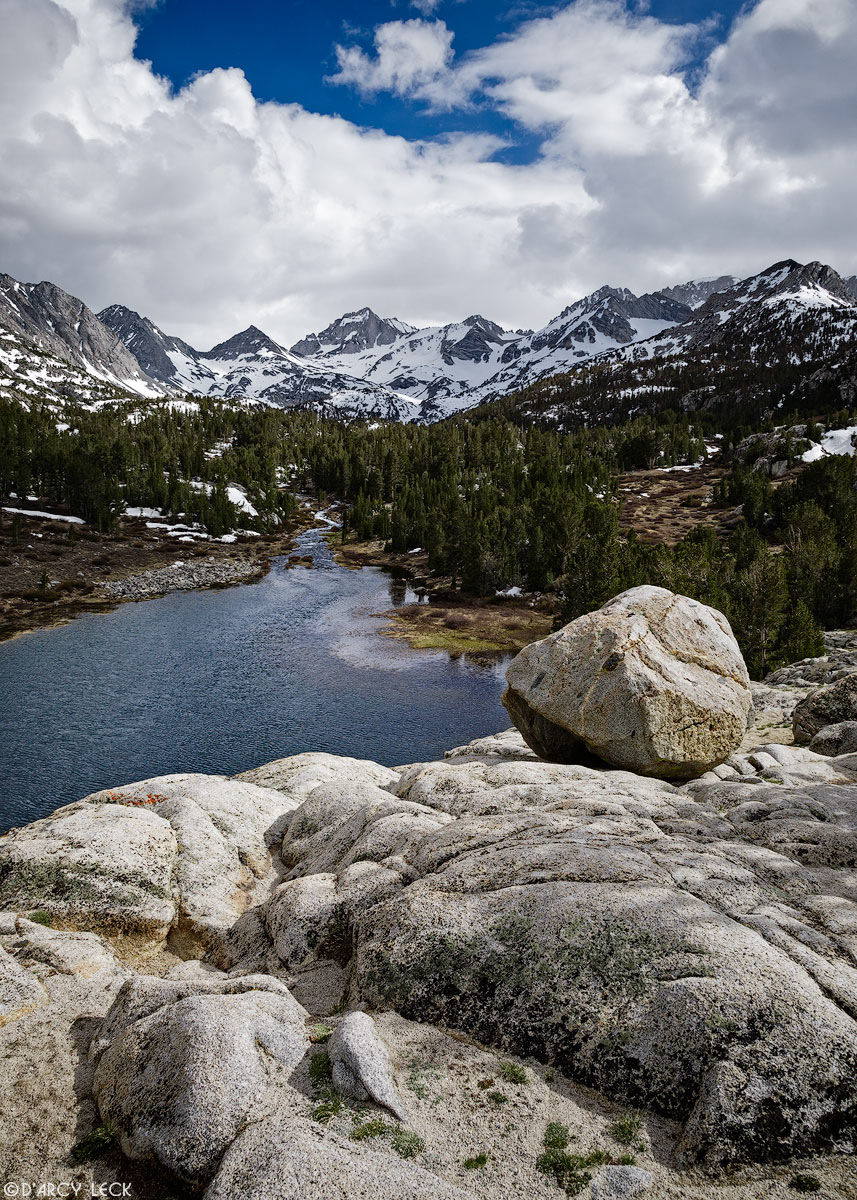 A rocky precipice overlooks Mack Lake with the Sierra Nevada Mountains beyond under late Spring snow showers in Little Lakes Valley, California
