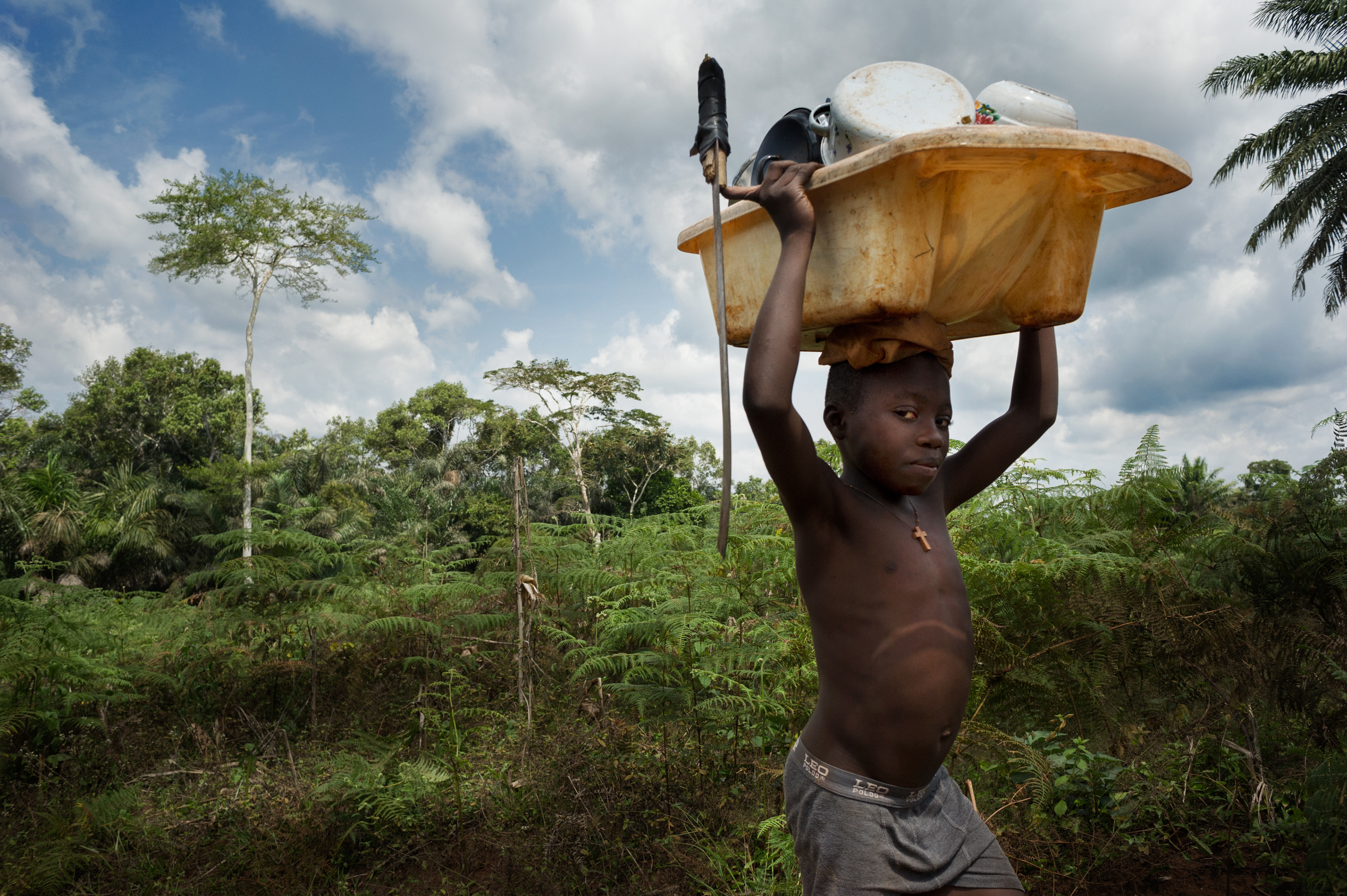 A boy returns from the forest, where he and his three siblings were washing dishes in a pond. He carries the dishes in an old baby bathtub. The machete he carries with him is the most common and versatile tool used by Cameroonians. It is useful in the forest for cutting leaves for dinner, firewood, and at home for preparing food. Abong-Mbang, Cameroon 2012