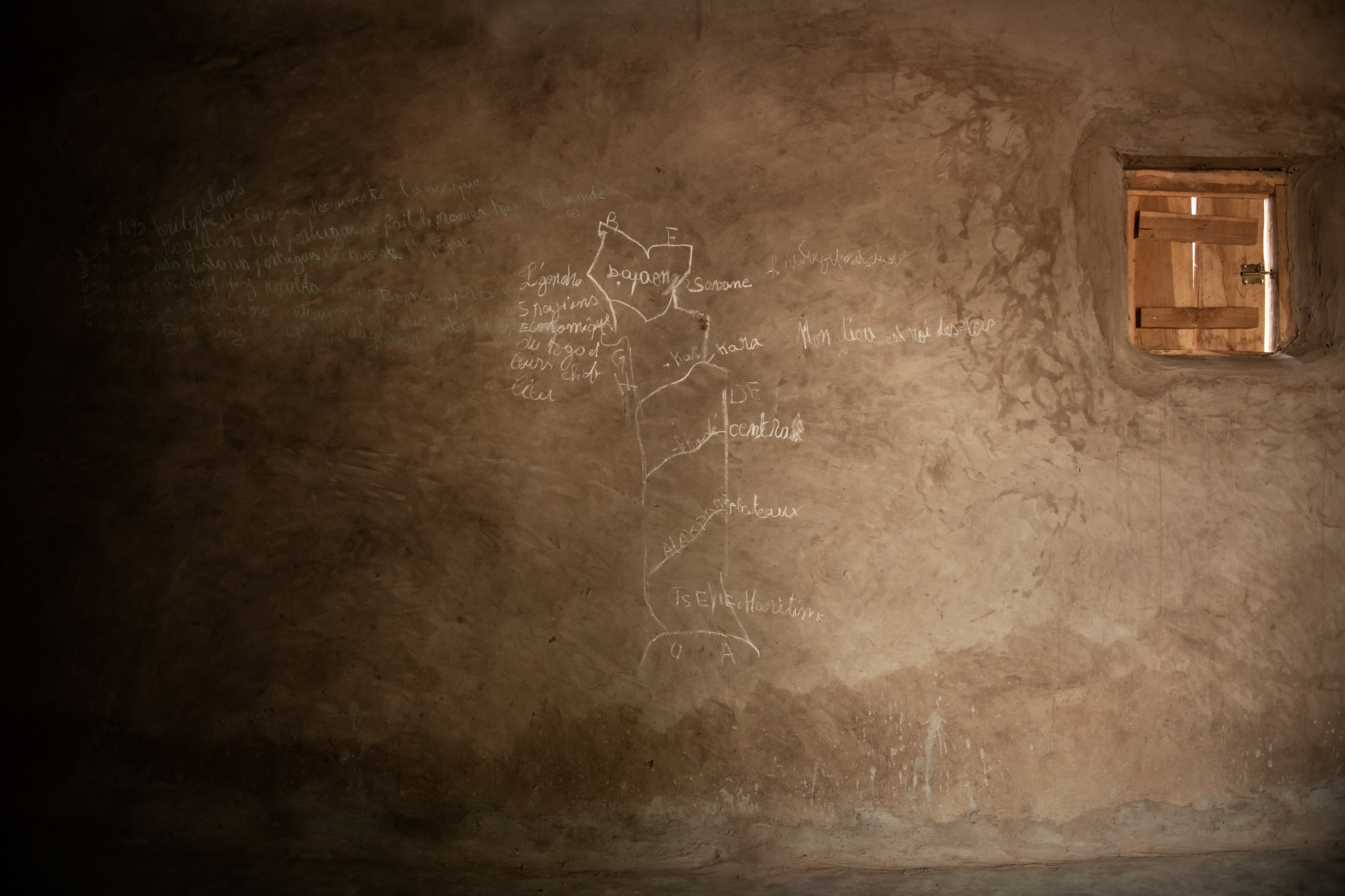 Lucy's bedroom, in Dilado, Togo. Lucy has 6 siblings. The room where she sleeps with her mother has no furniture or objects, not even a sleeping mat. On the wall is a chalk map of Togo divided into regions. Drawing with chalk on the wall is the most common way to learn at home. Children do not have notebooks, and at school they also use slates on which they write with chalk.  Togo 2023