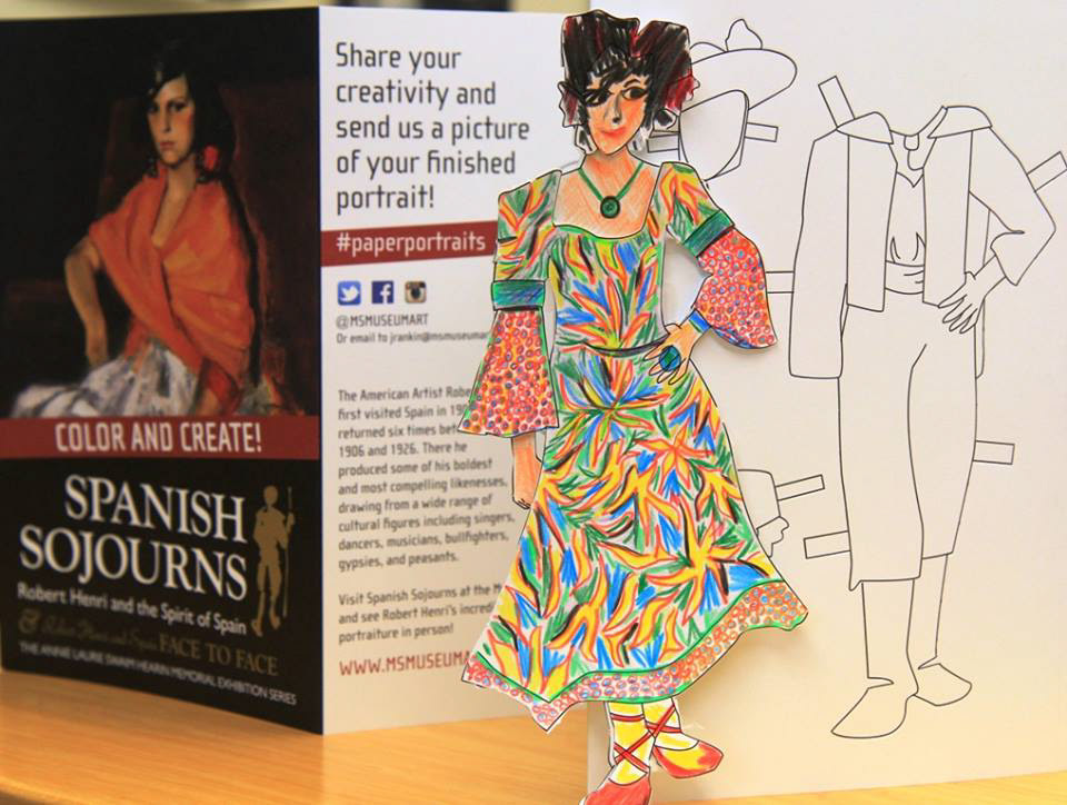 A brochure of a paper doll project standing on a wooden desk. In front of the brochure is a completed doll in a colorful dress.