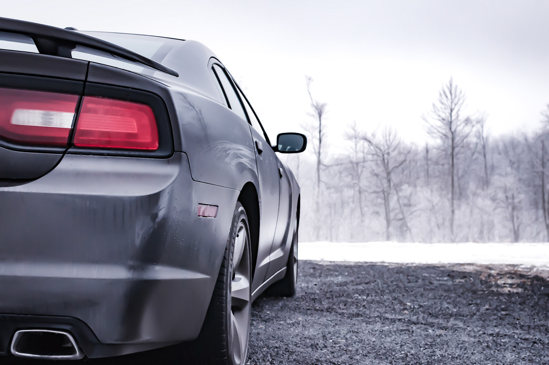 The image shows a close-up of the rear of a silver Dodge Charger parked on a snowy road. The car is parked on the side of the road, with snow blanketing the ground and the trees lining the road in the background.