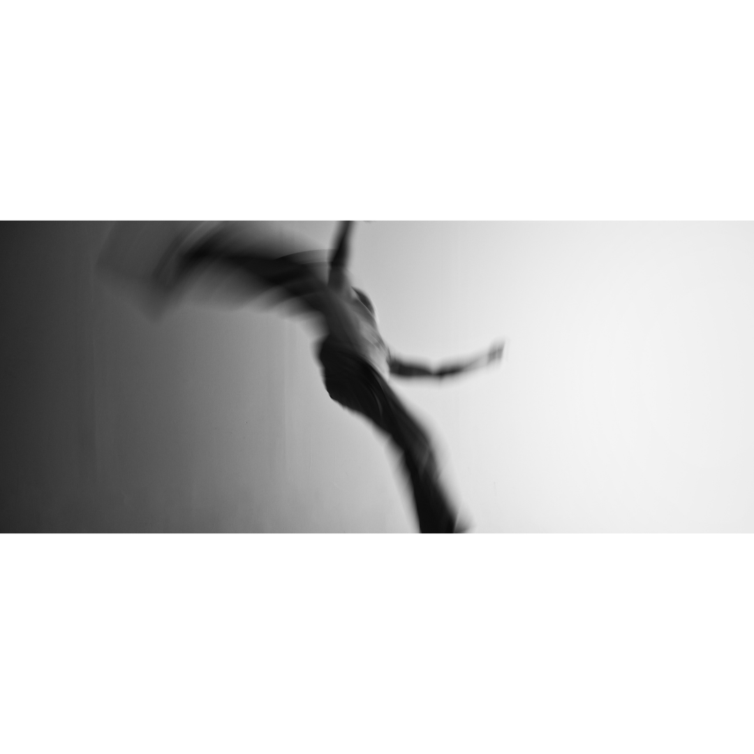 Black and white wide-format photograph of a figure suspended mid-leap, body dissolving into extreme motion blur against a light background, movement photography, MINUSELEVEN AUDIOVISUAL