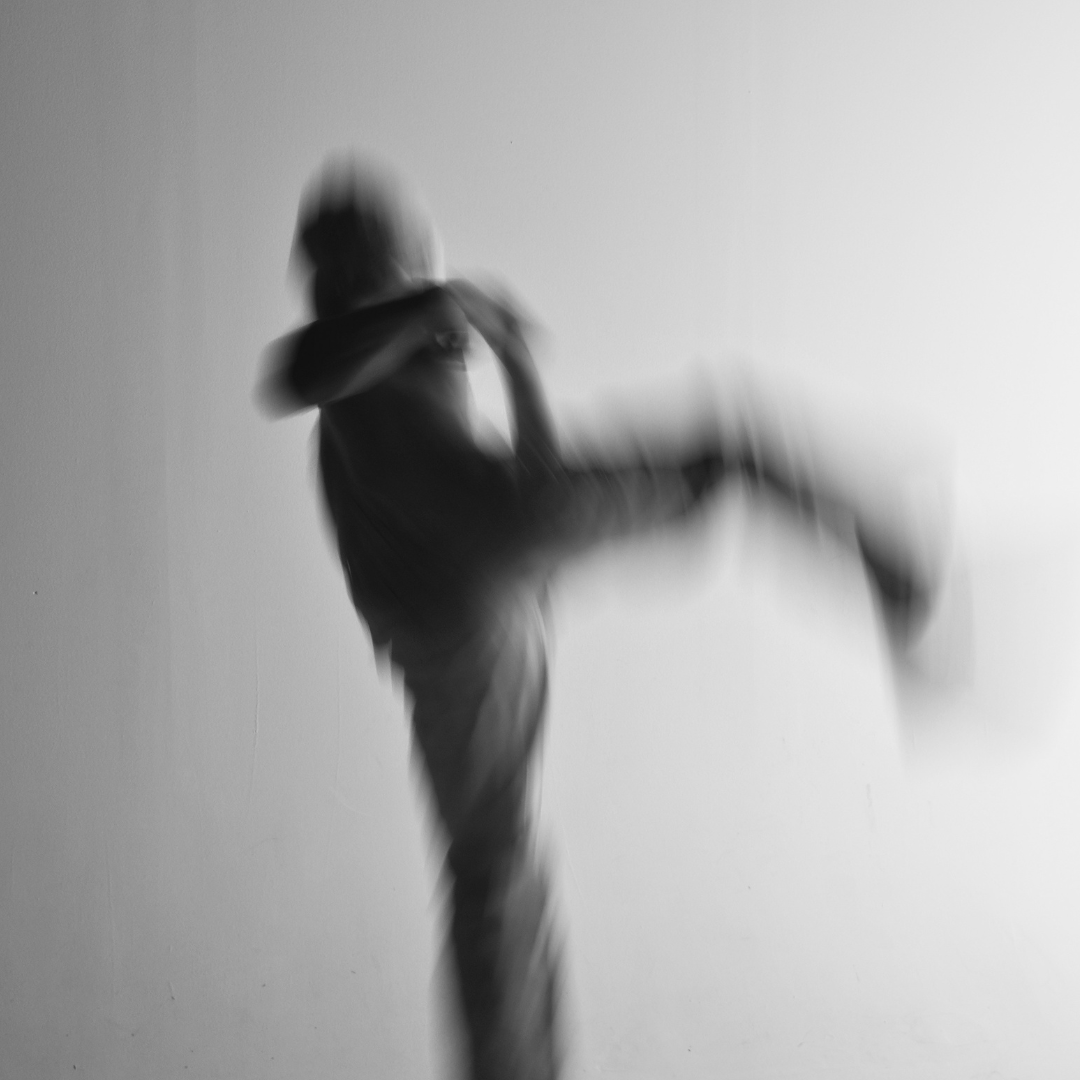 Black and white motion blur photograph of a masked figure performing an aerial high kick, martial arts movement photography, Bianchetto white jeans, MINUSELEVEN AUDIOVISUAL