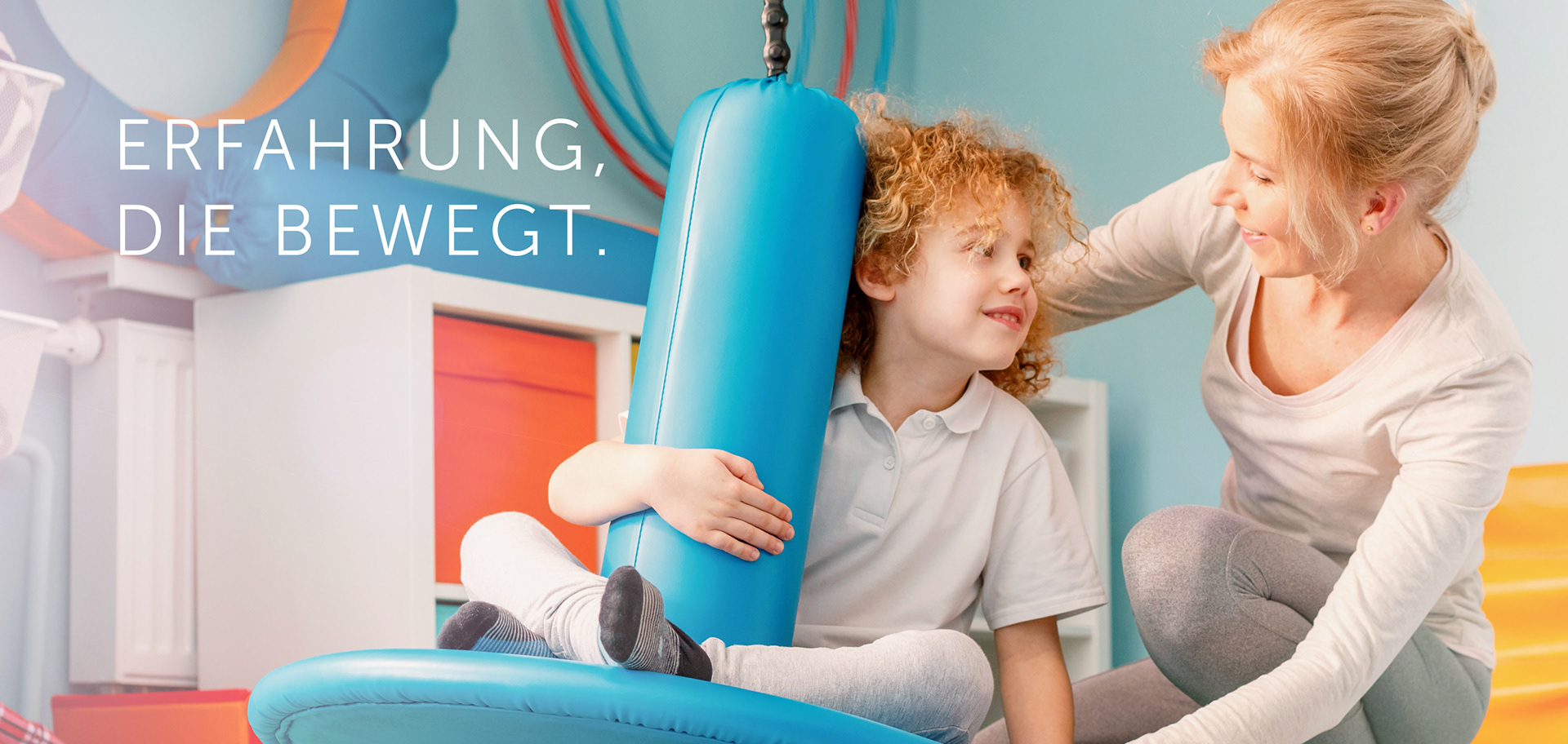 Eine Ergotherapeutin unterstützt ein fröhliches Kind auf einem blauen Therapiegerät in einem bunt eingerichteten Behandlungsraum – Momentaufnahme aus der Arbeit in der Ergotherapie Rosenthal. Claim: Erfahrung, die bewegt.