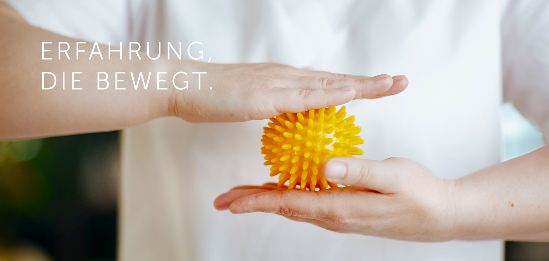 Zwei Hände halten einen gelben Igelball – eine Hand darunter, eine darüber – zur sensorischen Stimulation in der Ergotherapie. Die Person trägt ein weißes Oberteil. Links im Bild steht der Claim: „Erfahrung, die bewegt.“