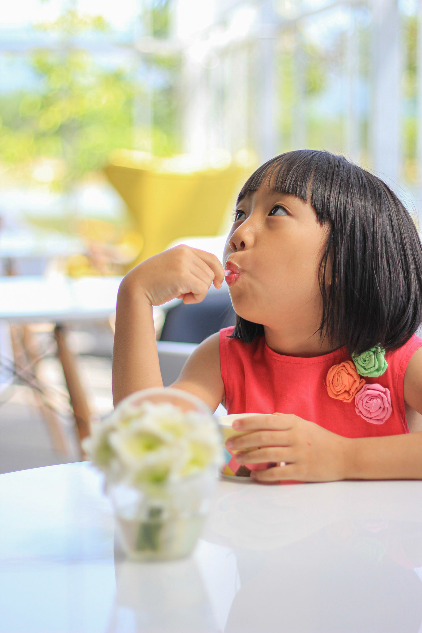 Asian girl wearing red dress is eating an ice-cream, looking up for something on the ceiling in a nice lighting interior. Focused on the girls's face.
