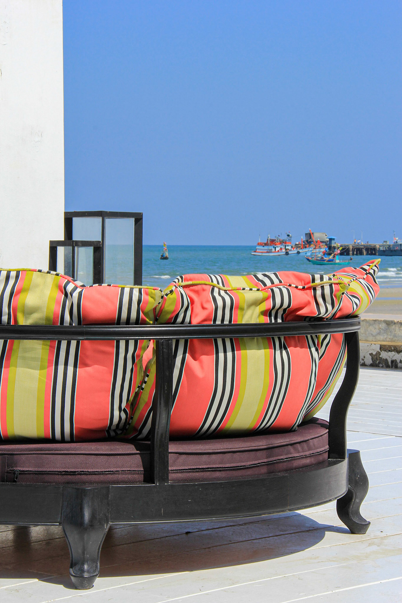Empty colourful sofa placed on white floor with an ocean view and far away fishing boat harbour.