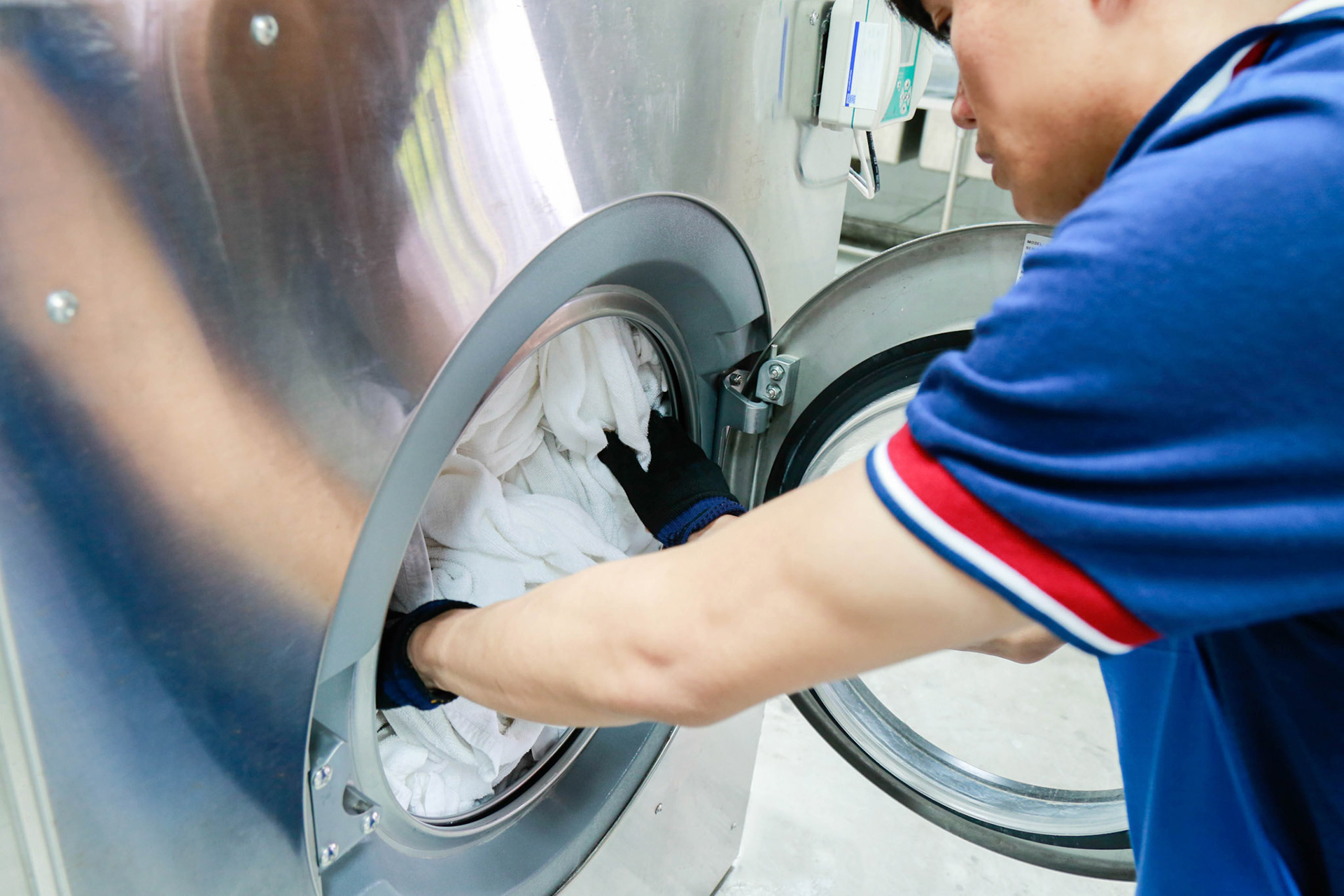 A laundry staff wearing a dark blue polo shirt is pushing towels into washing machine. Shot taken in the factory.