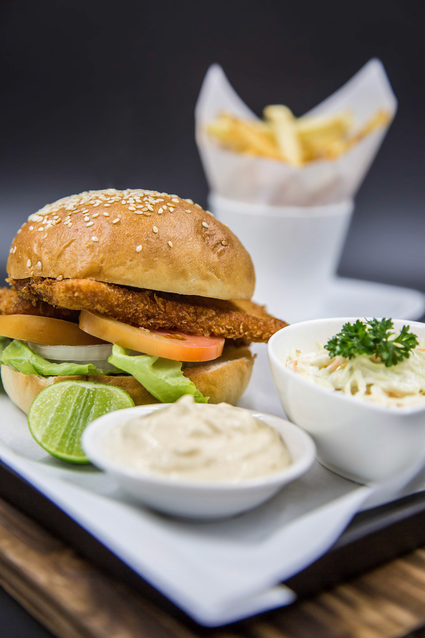 Fish burger set served with french fries and coleslaw, tartar sauce and lemon on the wooden tray with dark background. Focused on fried fish and vegetables.