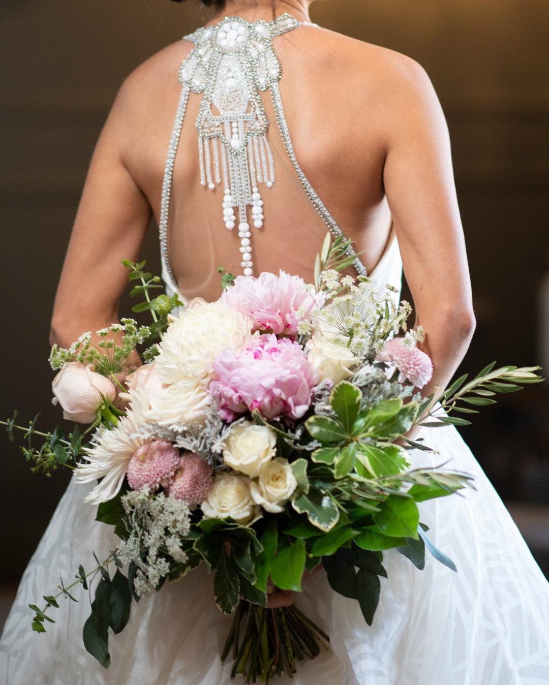 From the back, the exquisite beaded details of the bride's wedding gown sparkle above a sumptuous bouquet of pink and white peonies, roses, and other flowers and greenery