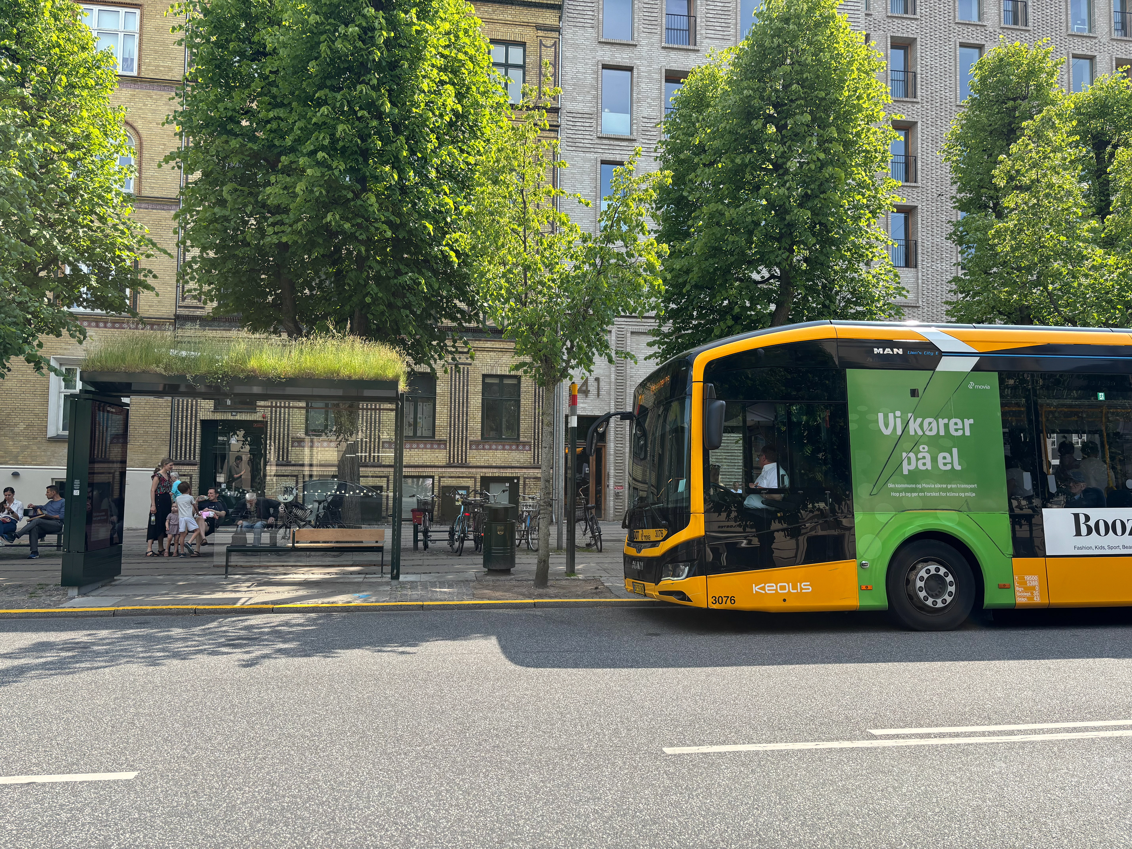 Electric city bus at a green-roofed bus stop with people waiting, trees, and buildings in the background.