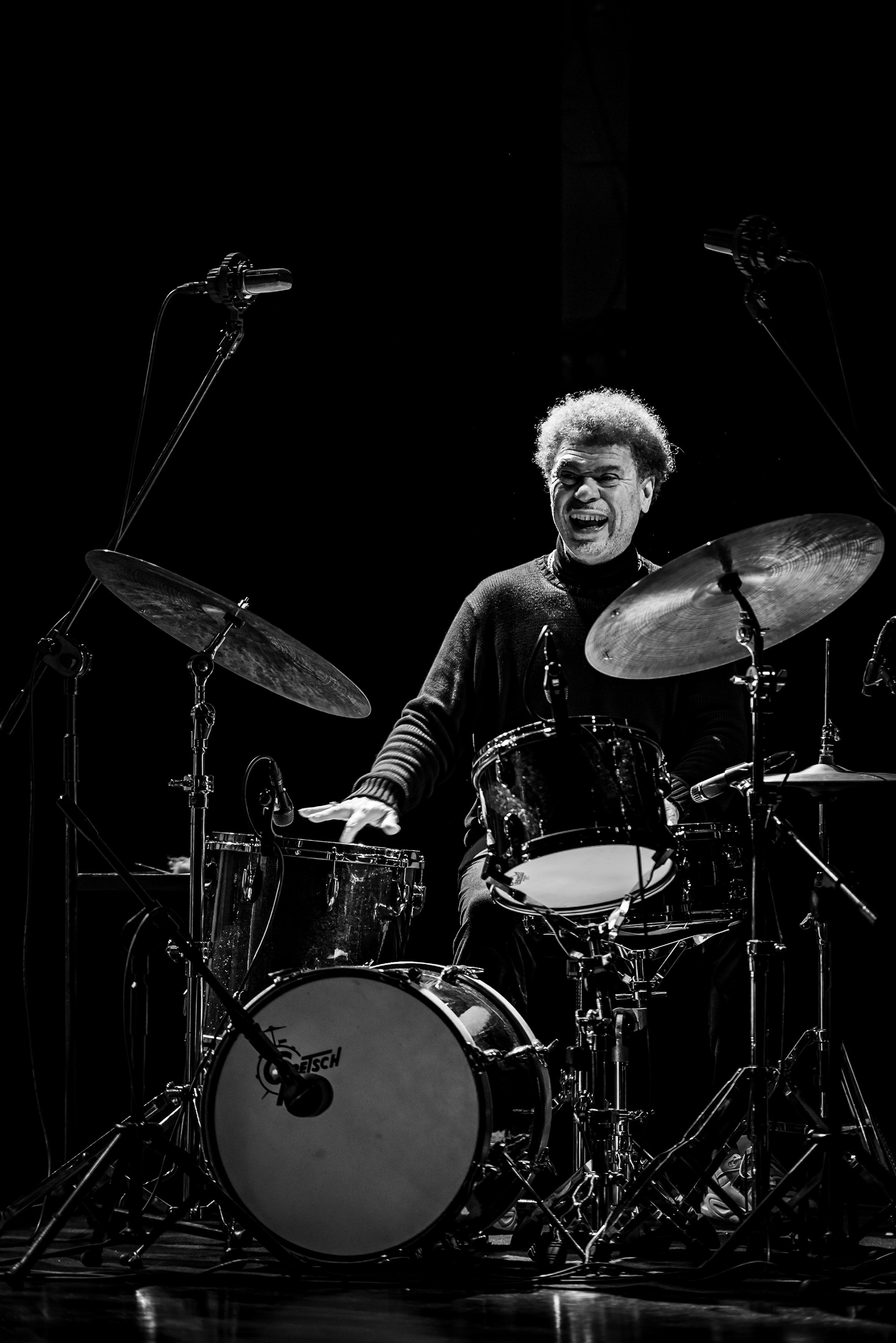 Drummer Lukmil Pérez performing in a haze of stage lights and deep shadows, dramatic black and white music photography, Szymon Ratajczyk Photography.