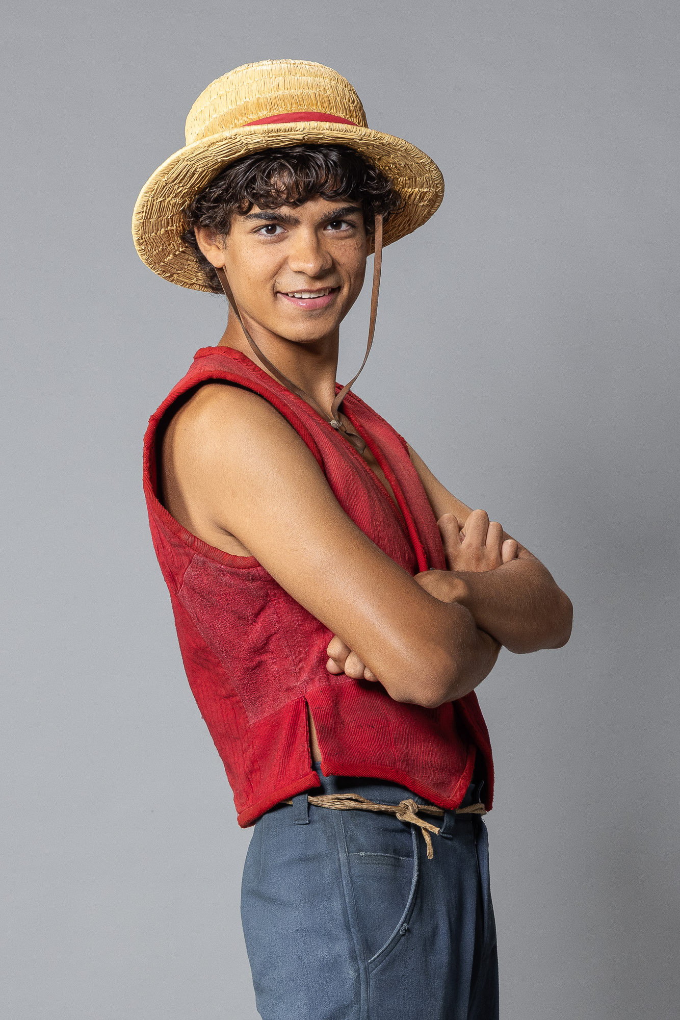 A professional studio portrait of Iñaki Godoy as Monkey D. Luffy for the Netflix live-action series 'One Piece,' captured by Principal Photographer Casey Crafford. The subject is shown from the waist up, wearing his iconic straw hat and red vest, posing with arms crossed against a neutral gray background. This high-resolution asset was commissioned to establish the definitive live-action visual for global marketing and brand partnerships