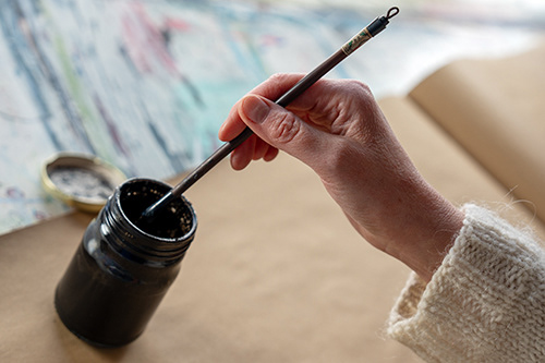 the artist ann-katrin persson dipping a pencil in a glass with black ink in her studio