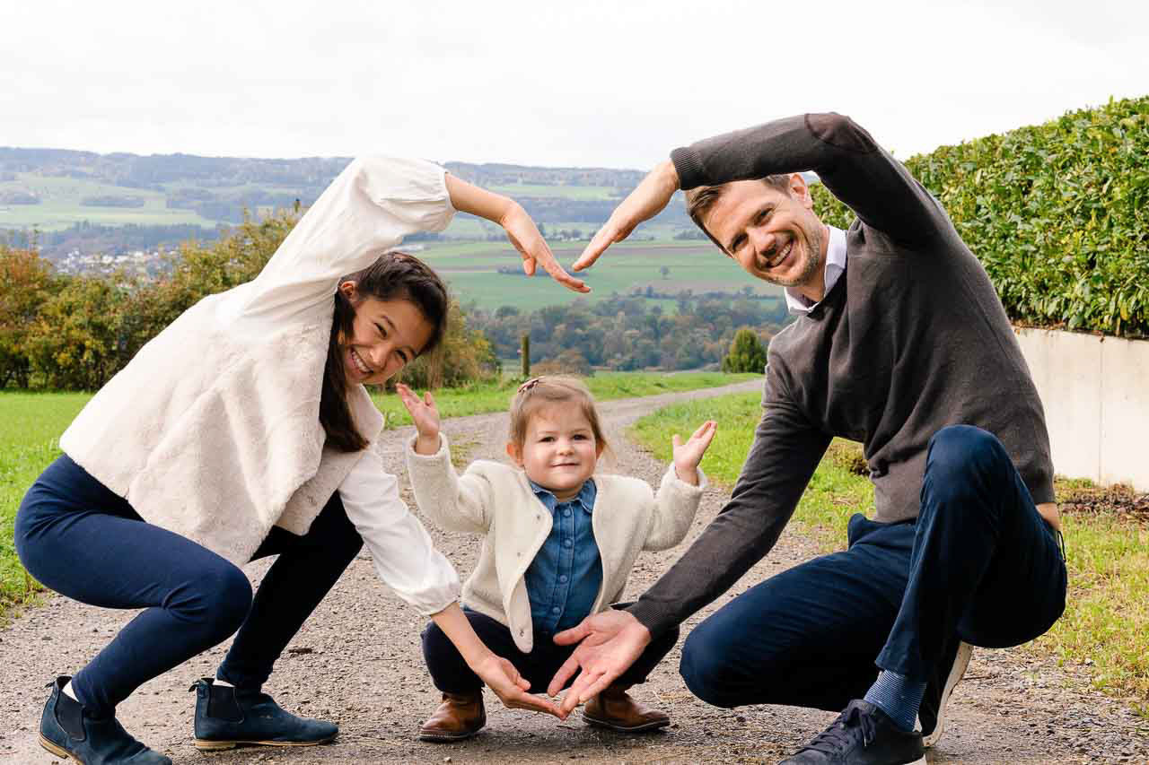 Vater und Tochter posieren mit Spass und Kreativität, während sie zusammen mit der grossen Schwester ein Herzsymbol bilden – eine Mischung aus Spiel und Emotion vor malerischer Landschaft.