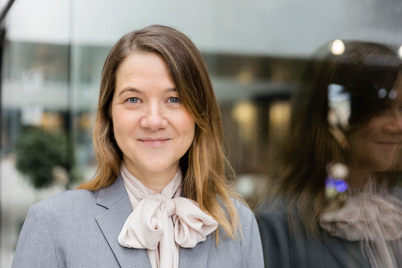 Professionelles Business-Porträt einer Frau in grauem Blazer, aufgenommen in natürlicher Umgebung mit reflektierendem Glas