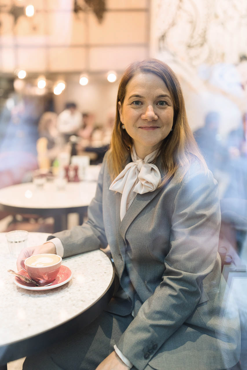 Professionelles Business-Porträt einer Frau in grauem Blazer, aufgenommen in natürlicher Umgebung mit reflektierendem Glas