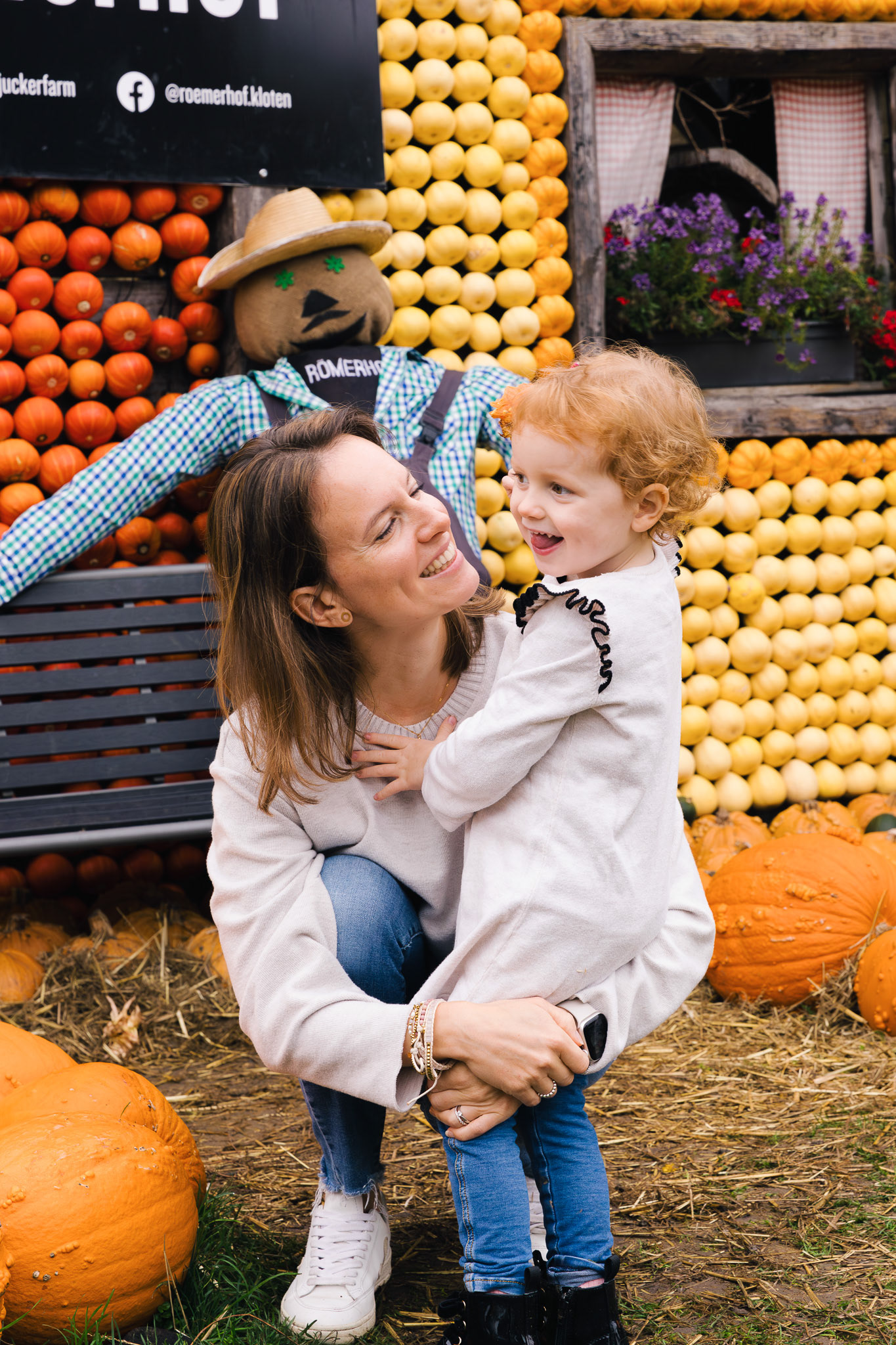 Eine Mutter hält ihre kleine Tochter liebevoll im Arm, vor einer Kürbiswand und herbstlicher Kulisse.