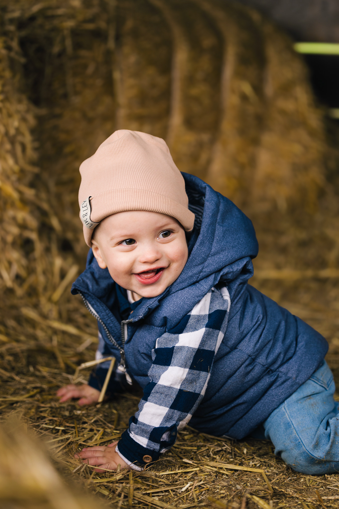 Herbstliches Babyshooting: Ein Baby mit beiger Mütze sitzt lachend auf Strohballen, ein Beispiel für natürliche Familienfotografie